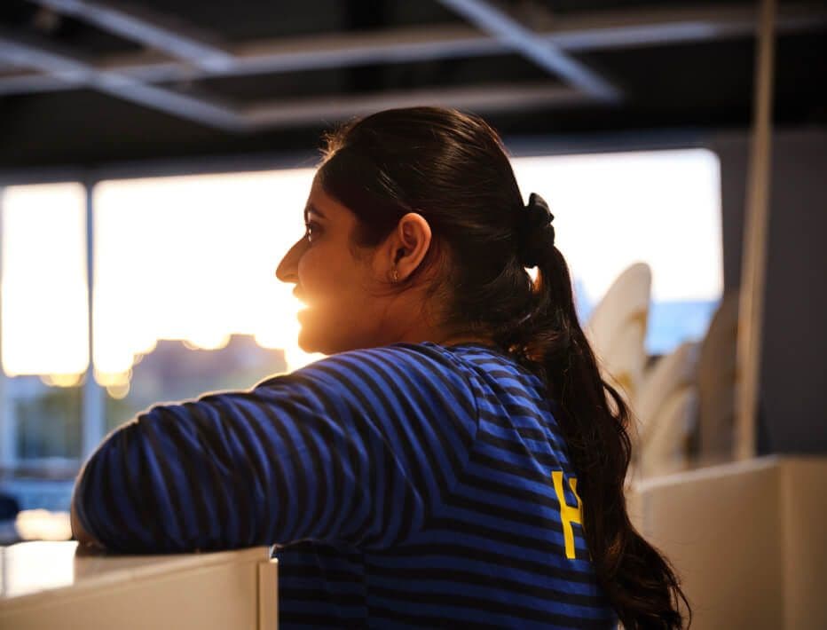 An IKEA co-worker leans against a piece of furniture. A sun sets in the window directly behind them.