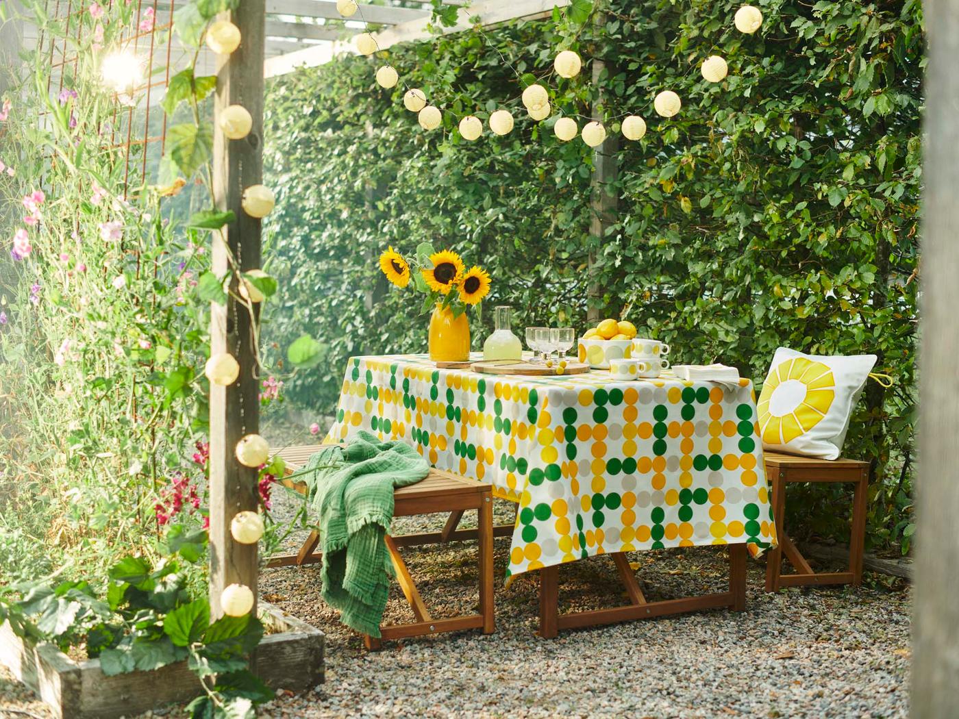 An outdoor space with NÄMMARÖ benches and a table with a BRÖGGAN tablecloth and glassware, and a vase of sunflowers.