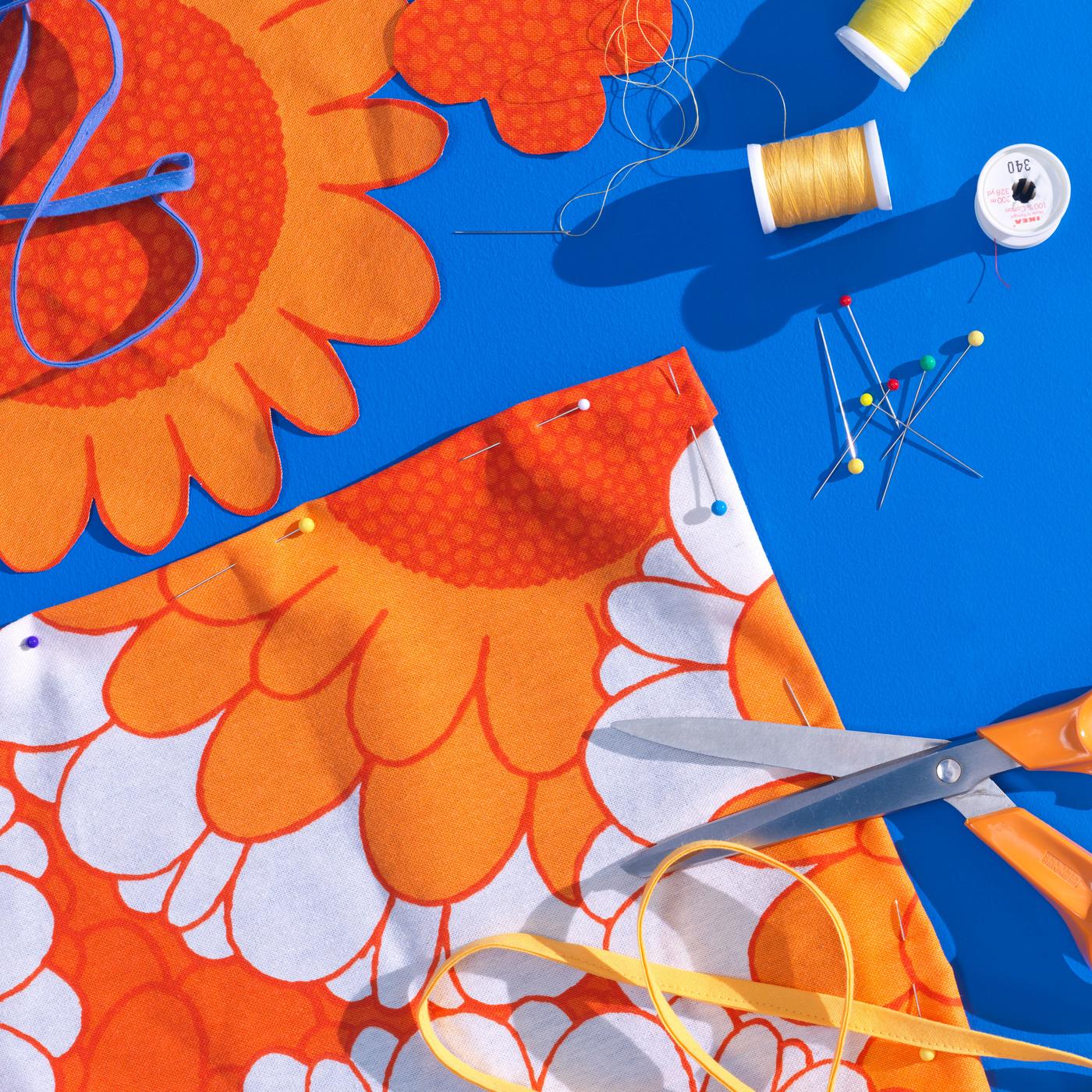 Photo of bright blue background with a colorful orange and white flower fabric. Next to it some yellow and orange yarn, scissors and needles.
