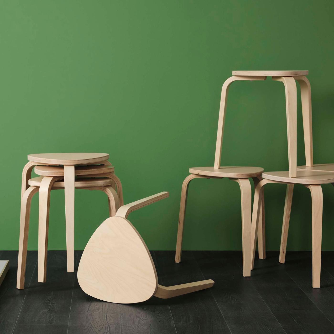 Seven KYRRE stools stand against a green backdrop. Some are stacked, one lays on its side and one is balanced on top of two other stools.