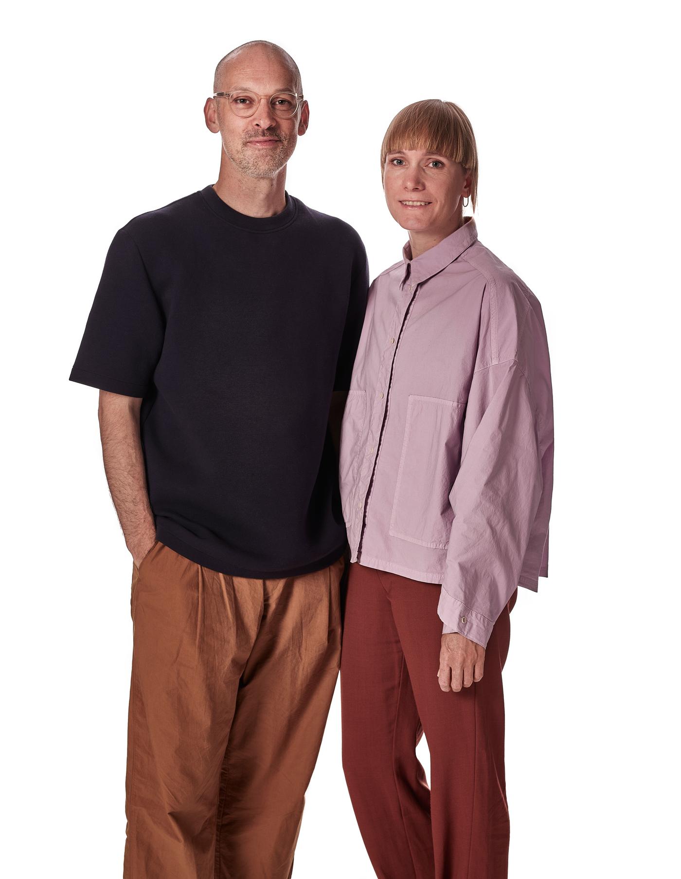 A portrait of a man with black t-shirt and brown pants to the left, standing next to a woman with pink overshirt and red pants.