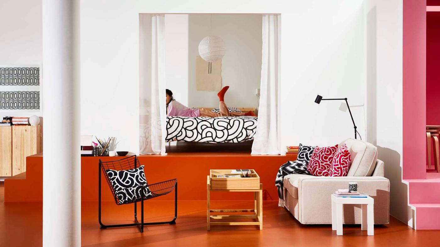 A room with a sofa, chair and table. In the background, a person is lying on the bed in the bedroom.