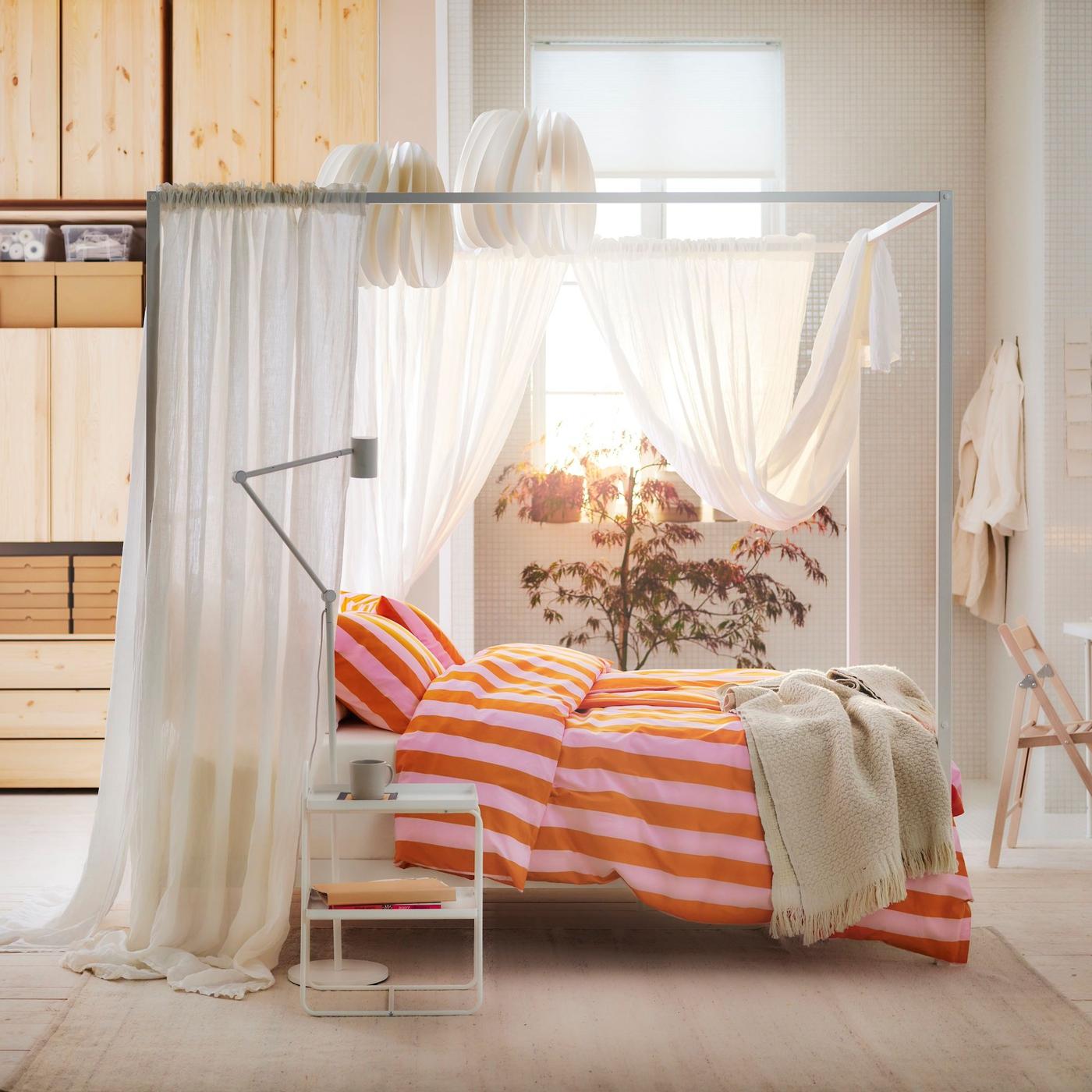 Warm light floods through the window of a bedroom with a VITARNA four-poster bed frame dressed with sheer white curtains and orange and pink-striped SLÅNHÖSTMAL bed linen.