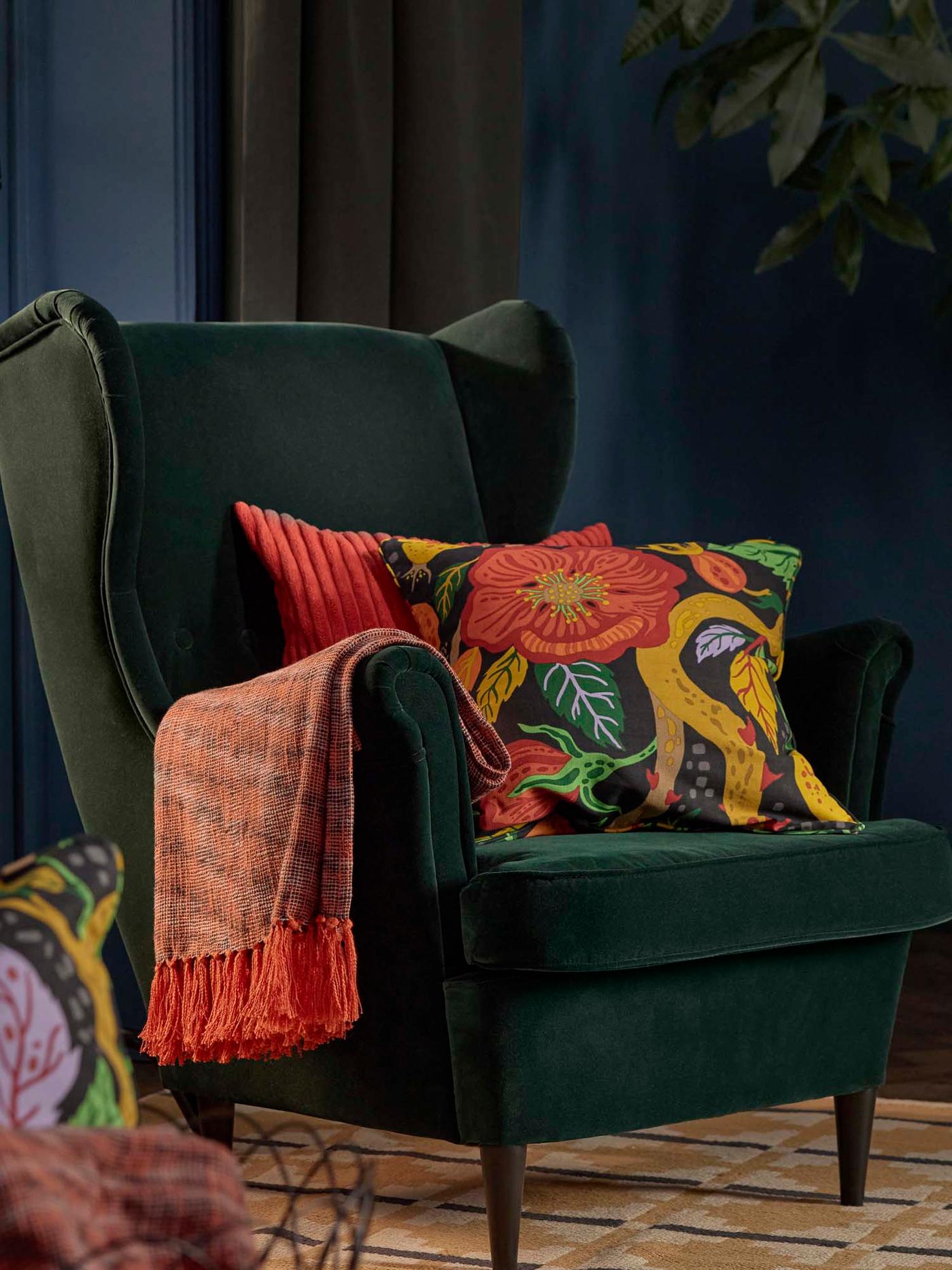 A room with an orange HÖSTAGILLE throw and two cushions in HÖSTAGILLE cushion covers on a dark green STRANDMON wing chair.