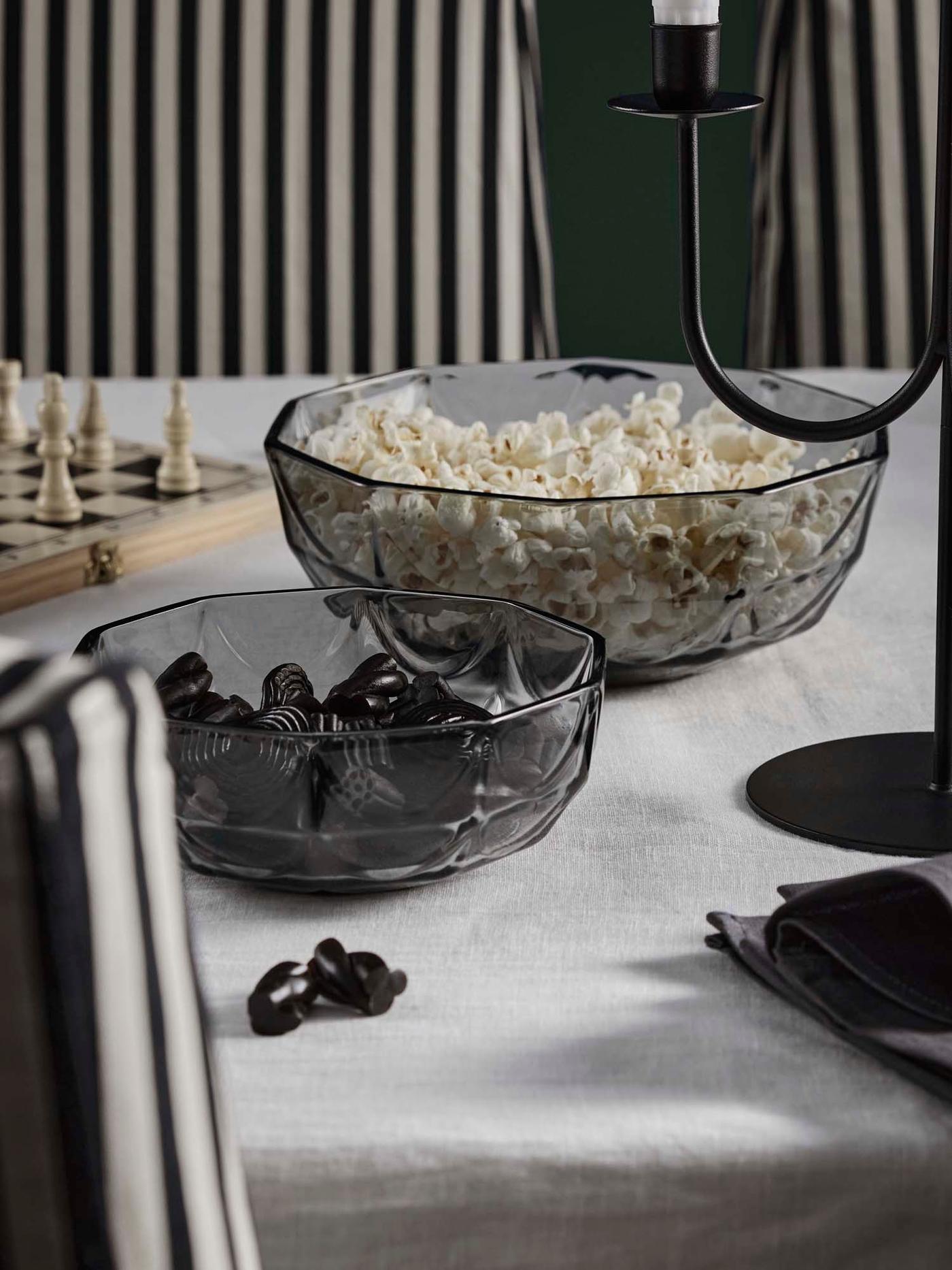 A table with two grey HÖSTAGILLE bowls filled with popcorn and liquorice atop a white KLYVBLAD tablecloth.