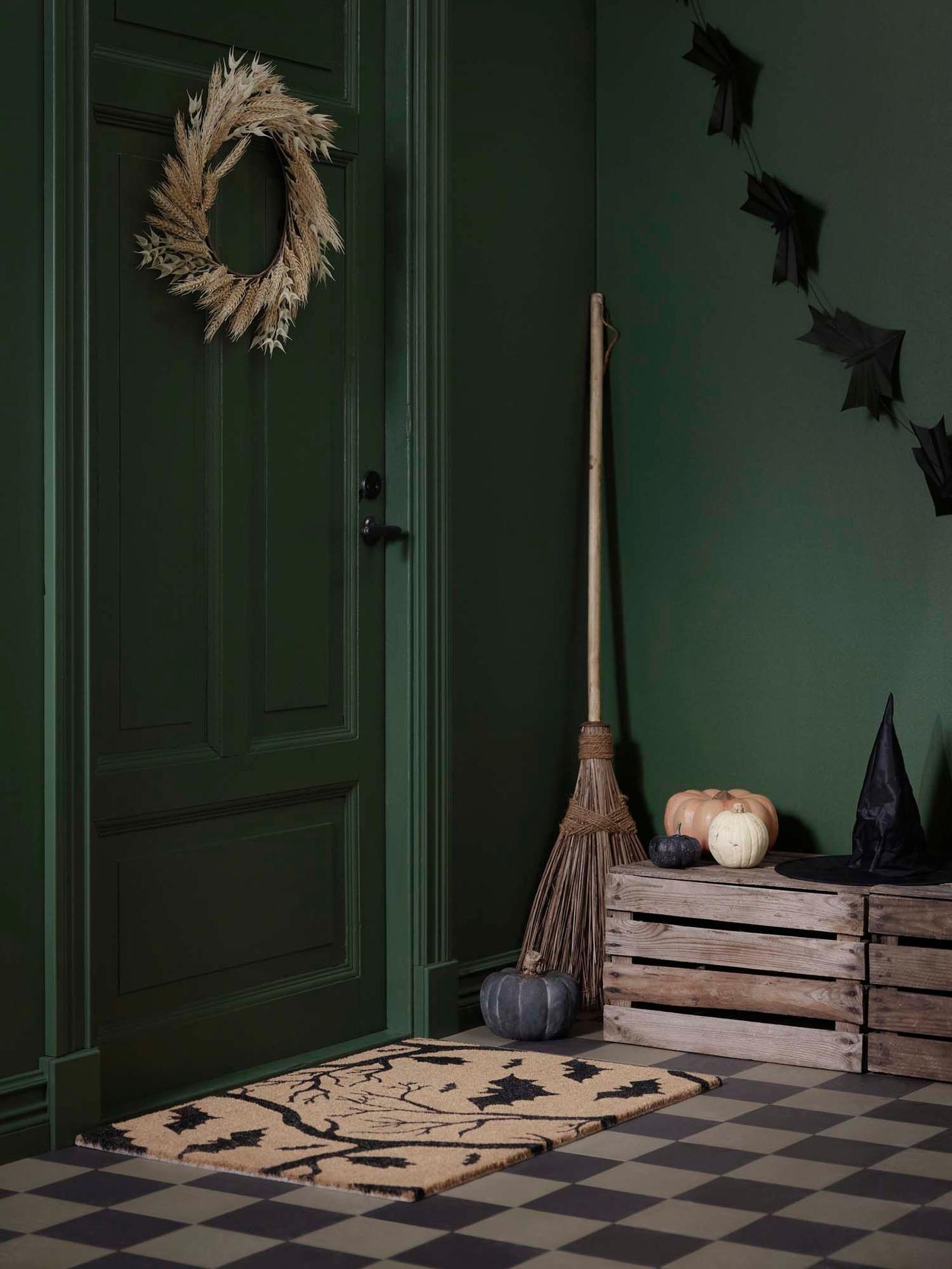 An entrance with a HÖSTAGILLE artificial wreath on a dark green door, and a HÖSTAGILLE door mat at the foot of the door.