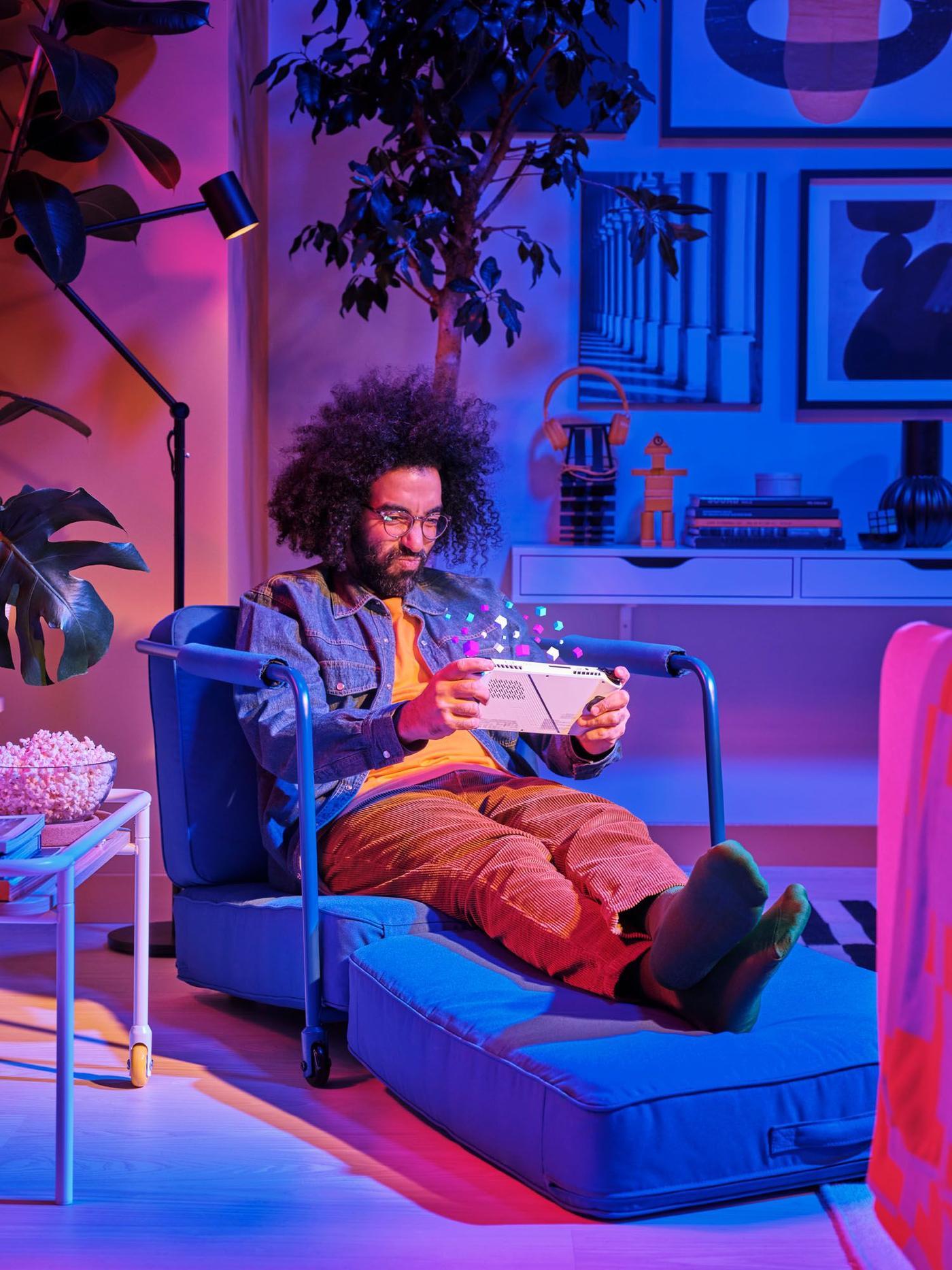 A person in a blue and pink-lit living area sits in a folded out BRÄNNBOLL gaming lounge chair playing a hand-held video game.