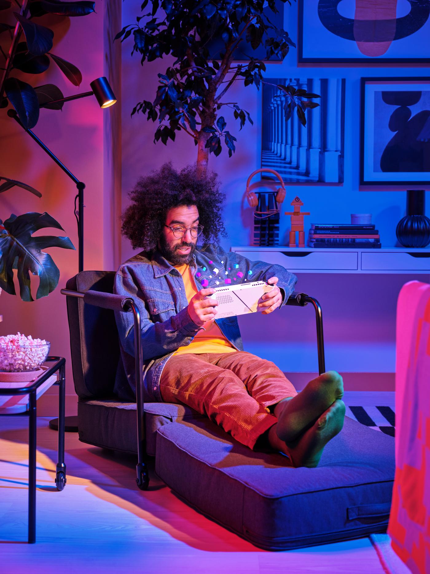 A man is sitting on a blue chair in a dark room, playing video games