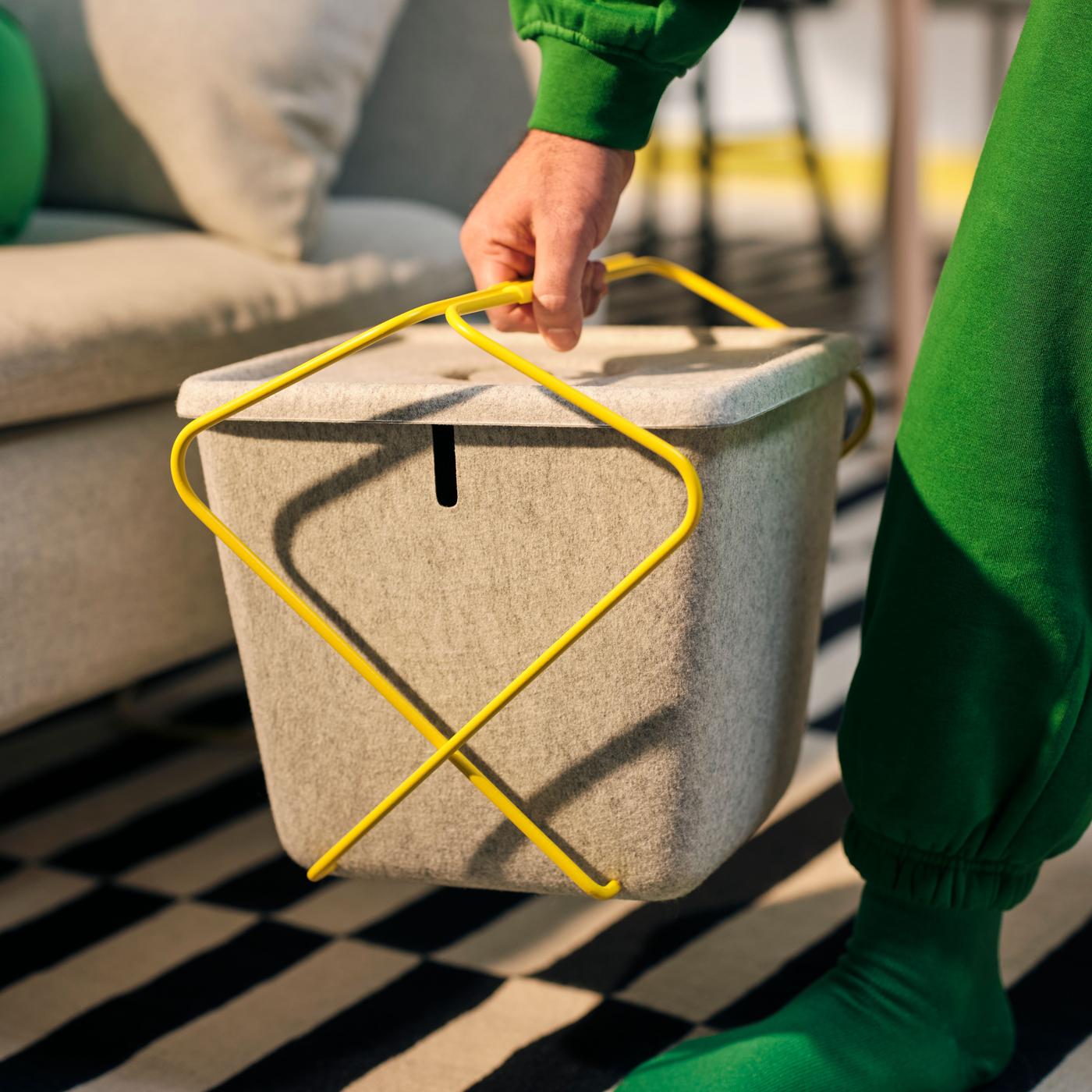 A hand is holding a beige and gray basket with yellow steel handles, carrying it.