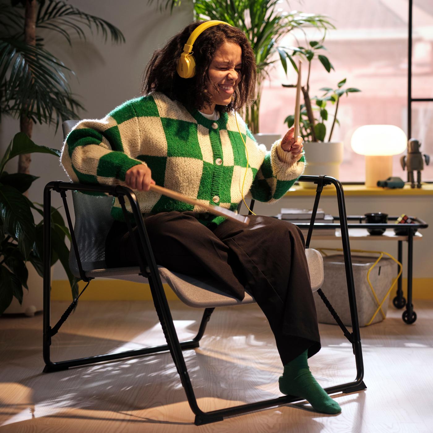 A girl with yellow headphones is air drumming with drumsticks, sitting in a swinging black easy chair