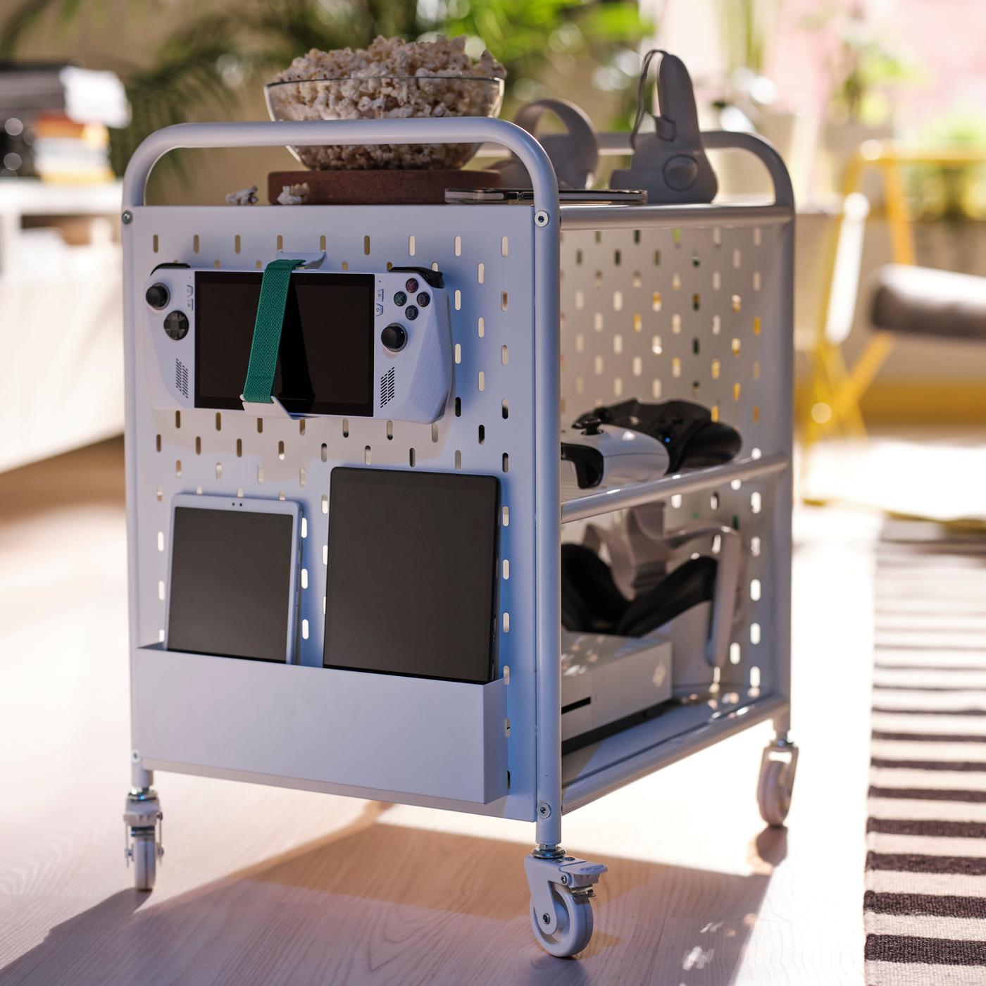 A white trolley with wheels is standing in the middle of the room, with video game controls, tablets and chords strapped on it.