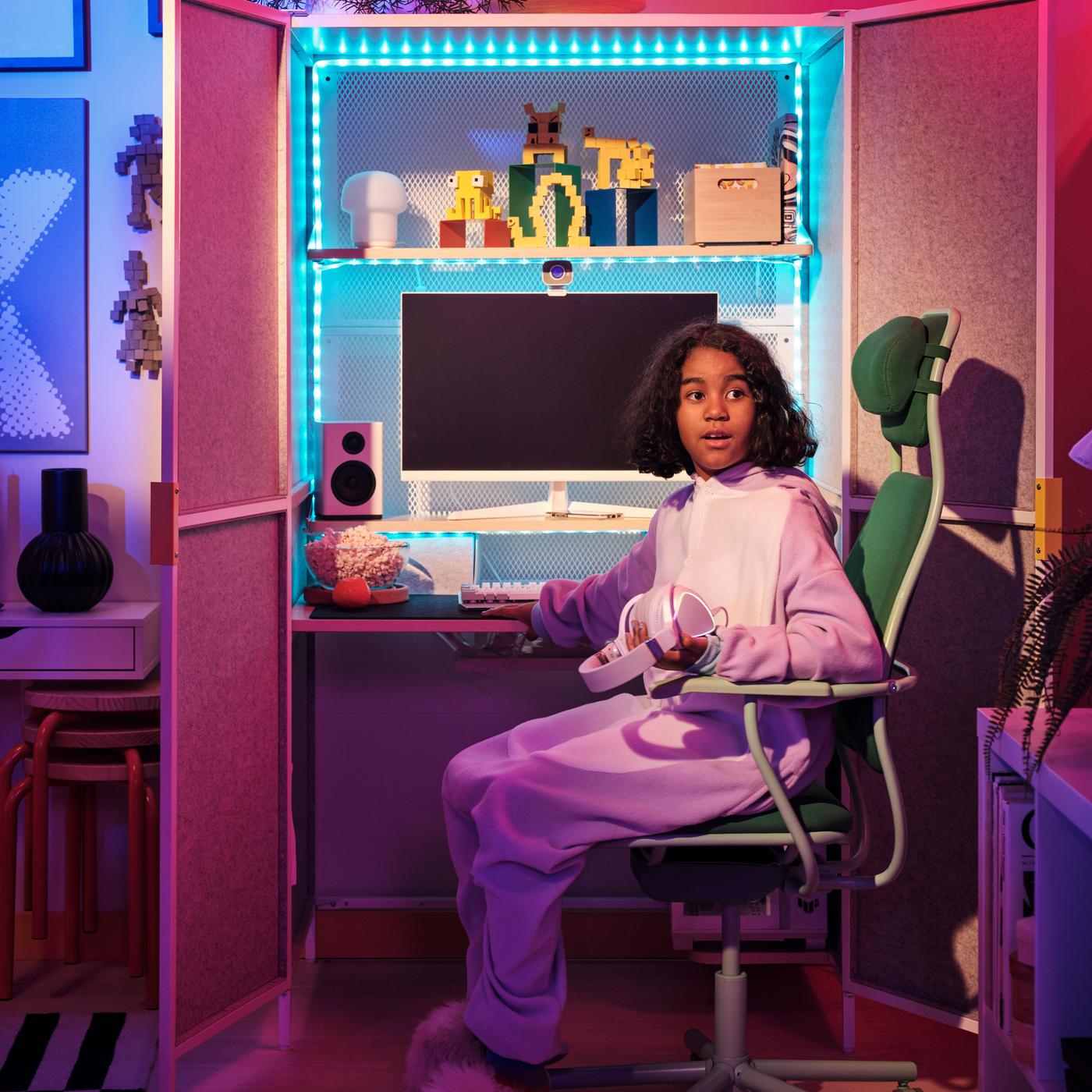 A girl is sitting in a green chair with a cabinet/gaming station with blue LED lights and filled with gaming equipment.