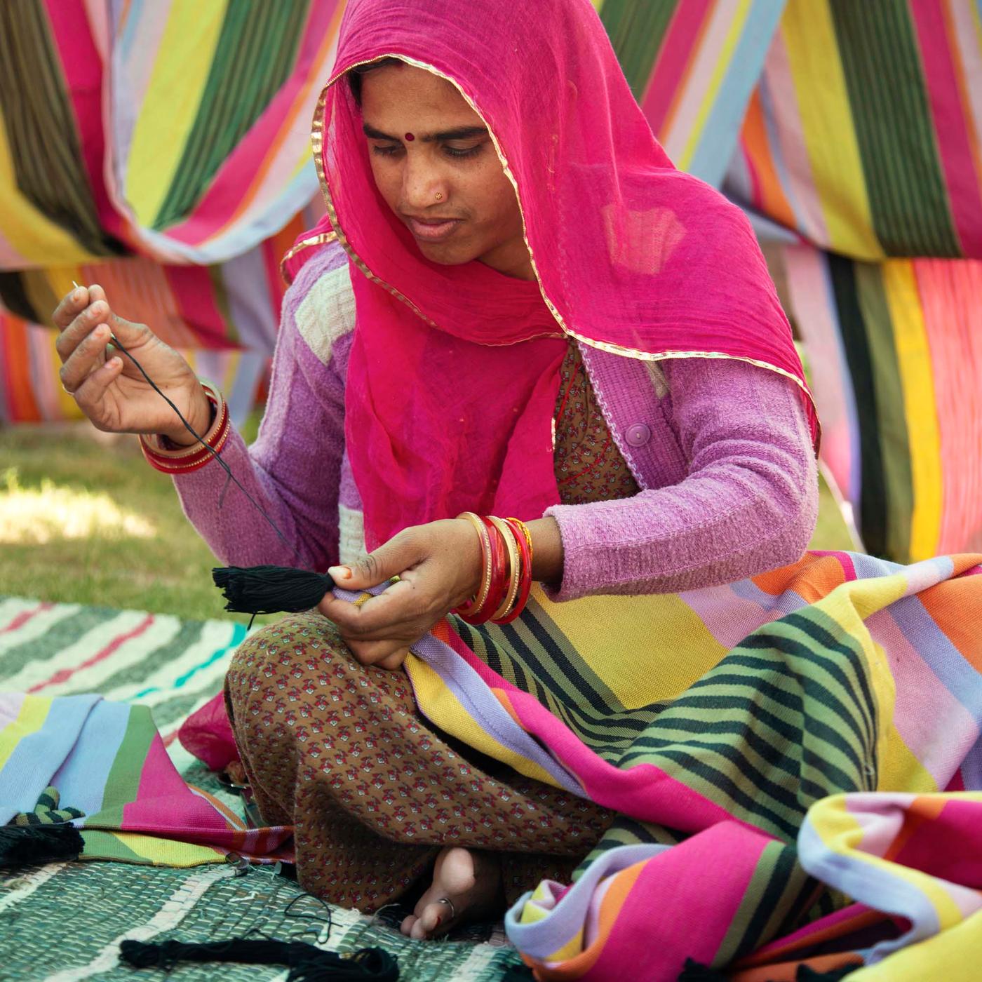 A person sits crosslegged while stitching a striped multicolour MÄVINN throw.