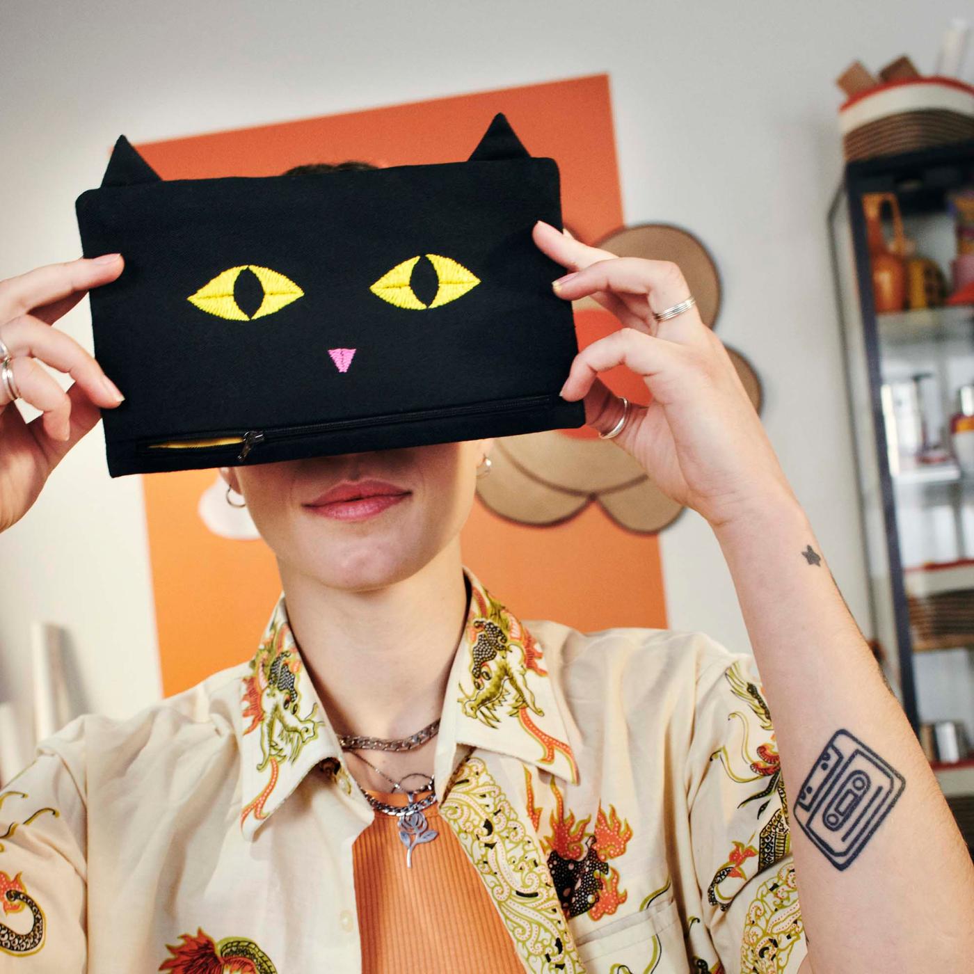 A woman holds a black bag with a cat face (from the MÄVINN collection) over her eyes, so she looks like she has cat eyes.