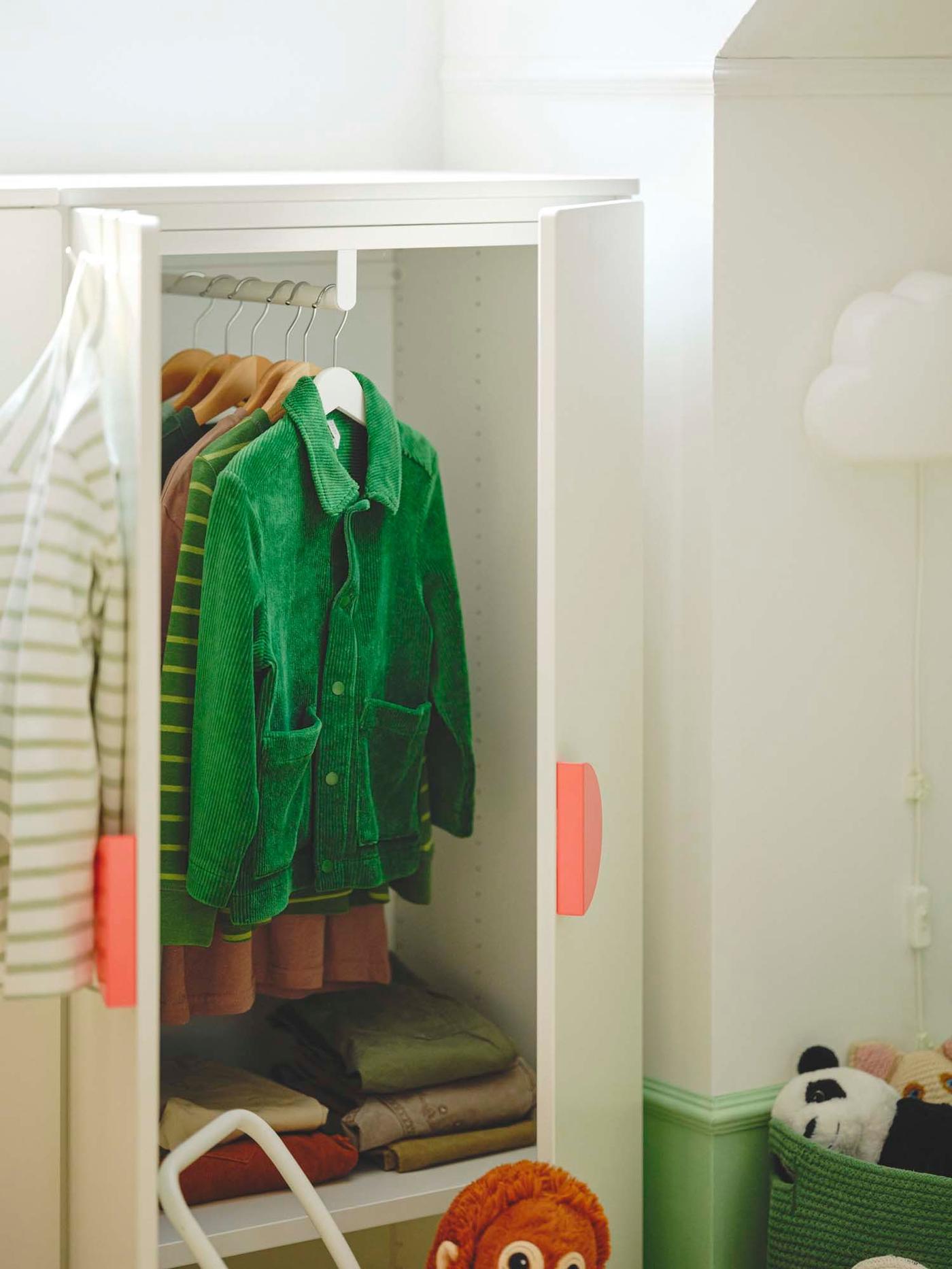 An open white wardrobe with pink handles reveals a green jacket and other clothes hanging inside.