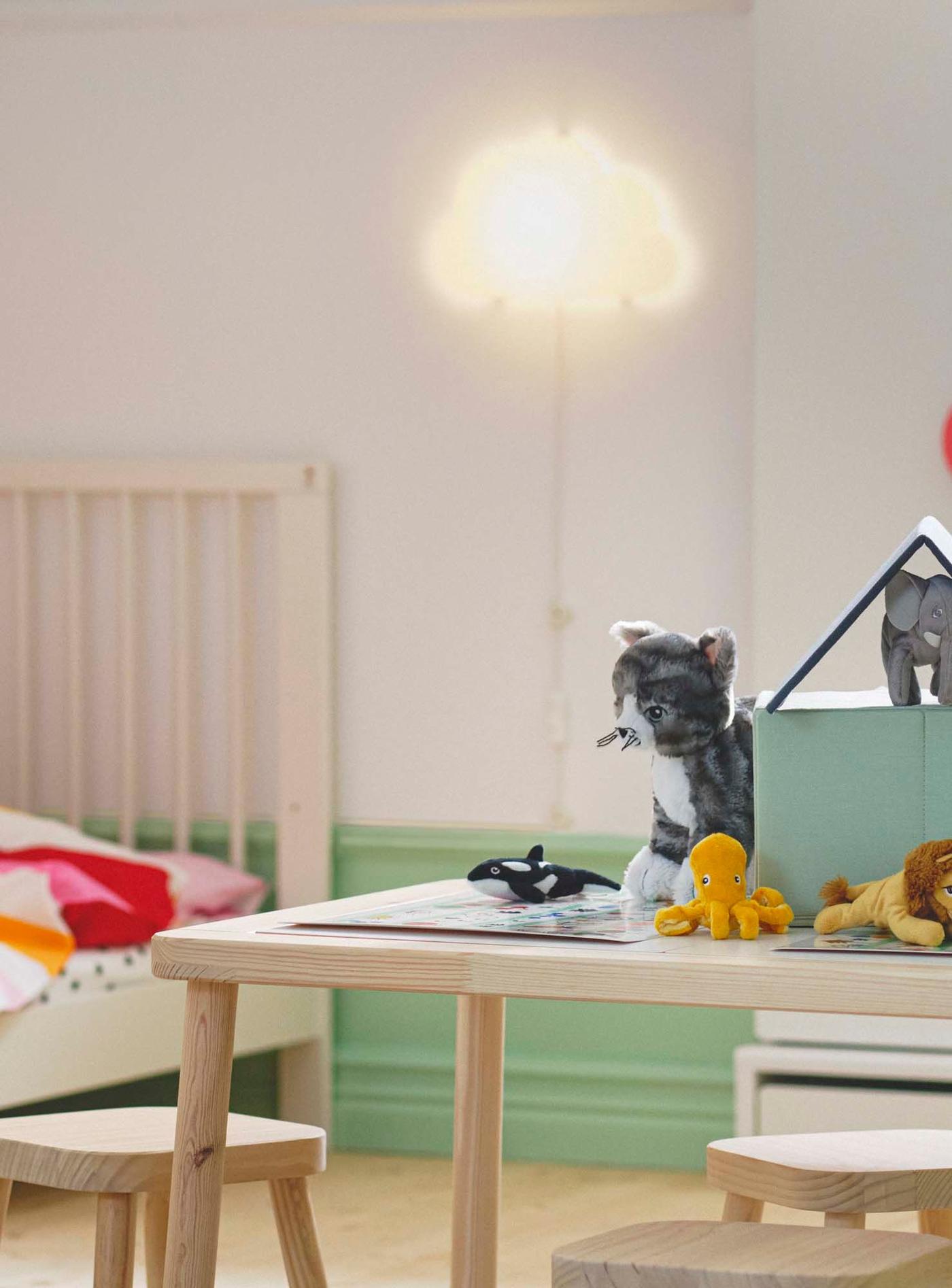 A white, illuminated cloud-shaped lamp hangs on the wall, above a white cot. A table filled with toys stands in the forefront.
