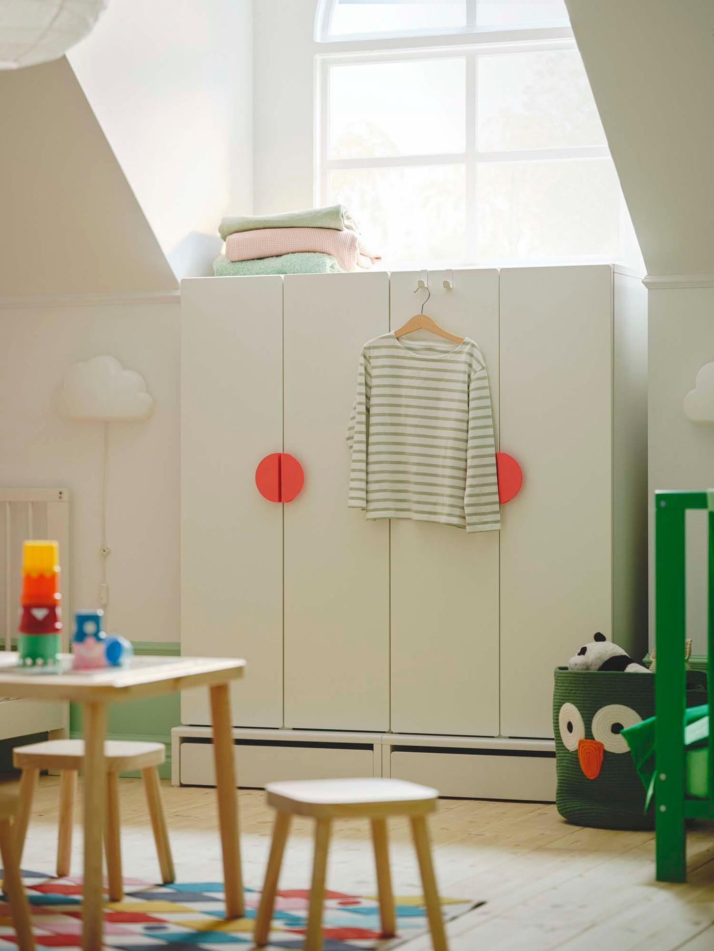 In a toddler's room, a white wardrobe with pink circular handles stands below a window. In front is a table with chairs.