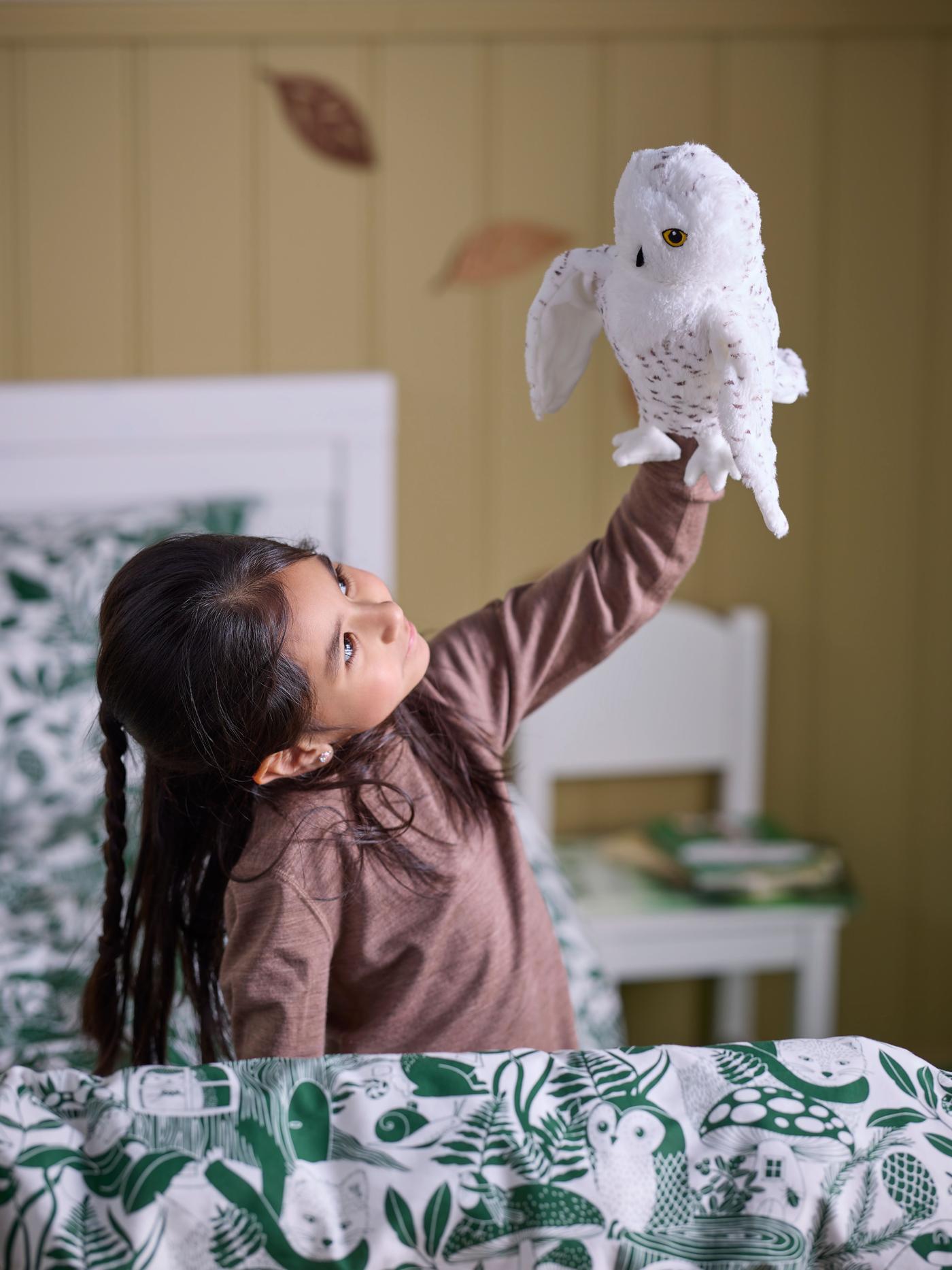A child sitting on her bed with SKOGSDUVA duvet covers. In her left hand show's holding a soft toys owl high up while looking at it.