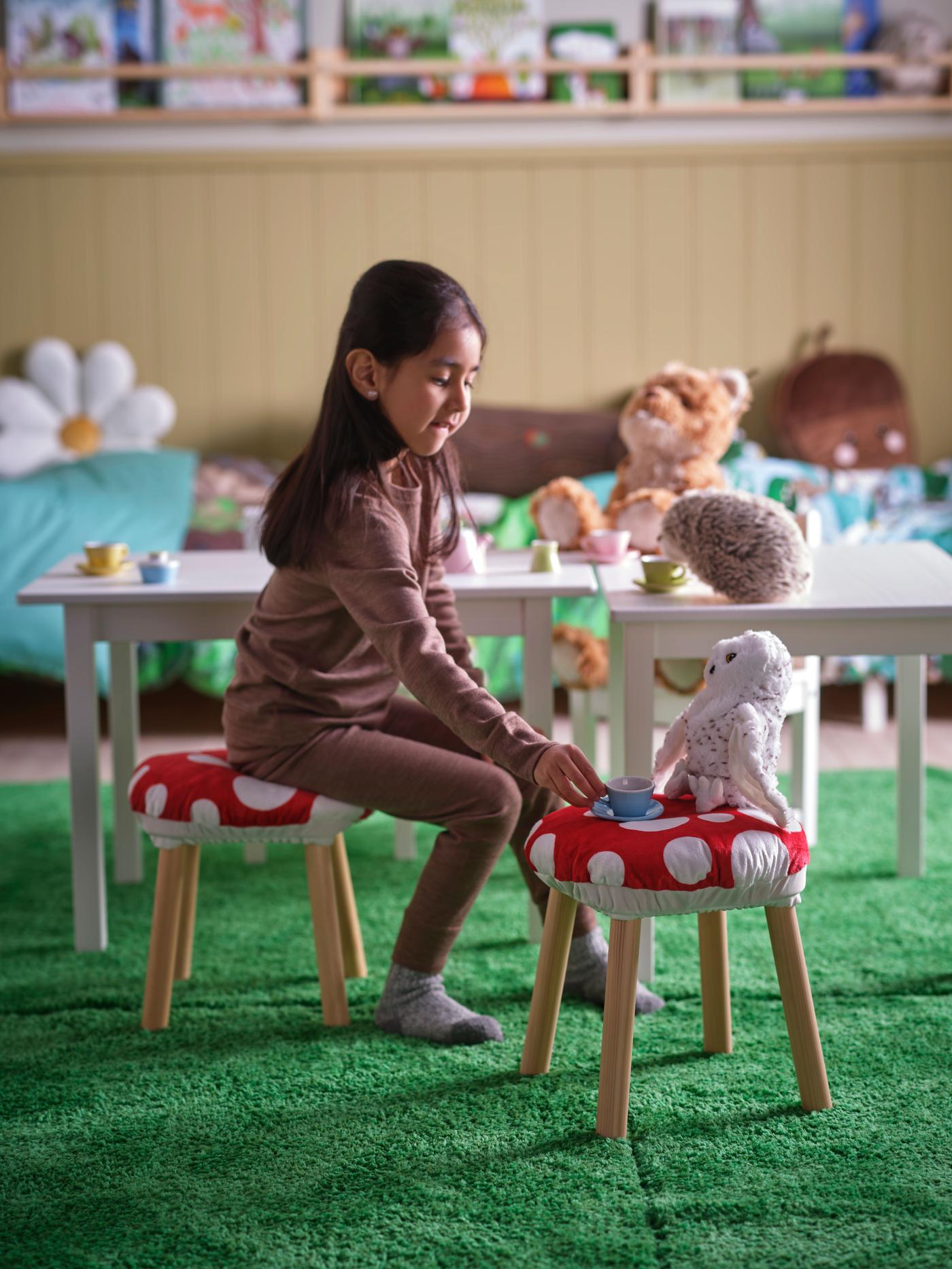 A child in a room surrounded by different products of the SKOGSDUVA collection. She is sitting on a small table surrounded by red and white toadstools. On one of the toadstools sits a soft toy owl that the child is offering a cup of tea.