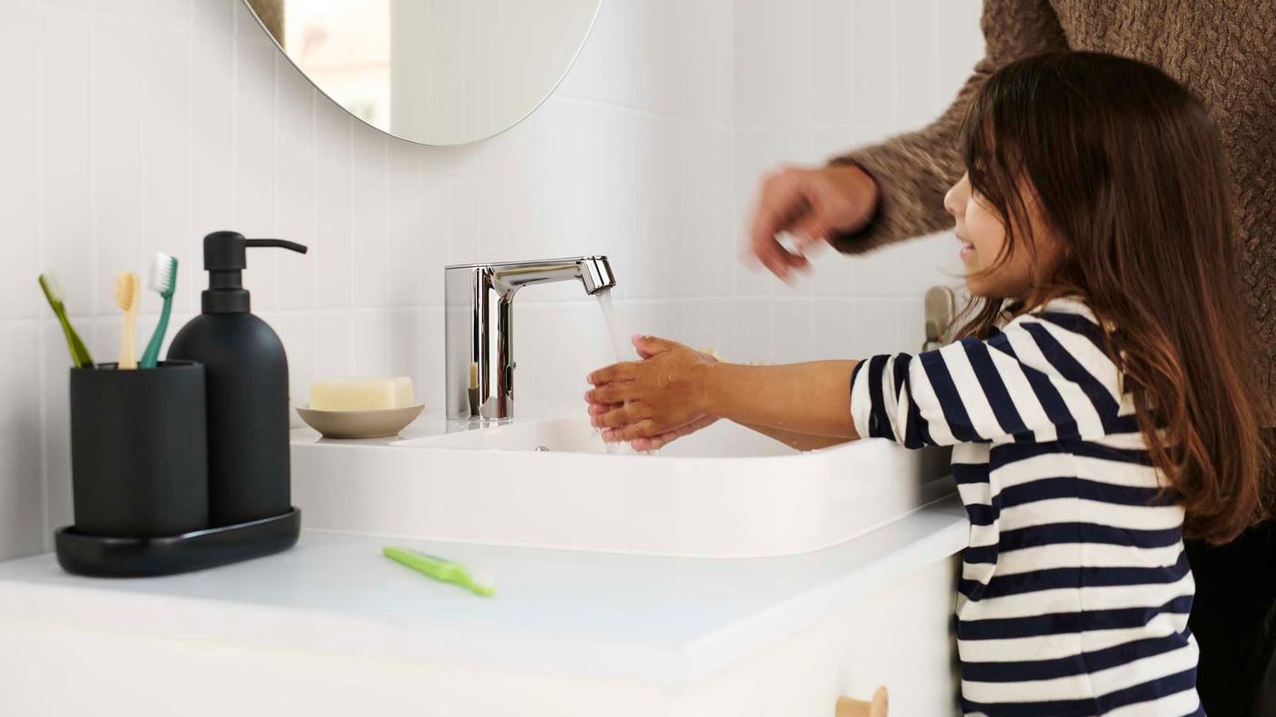 A child washing their hands under a chrome-plated BROGRUND wash basin tap with sensor in a white bathroom.
