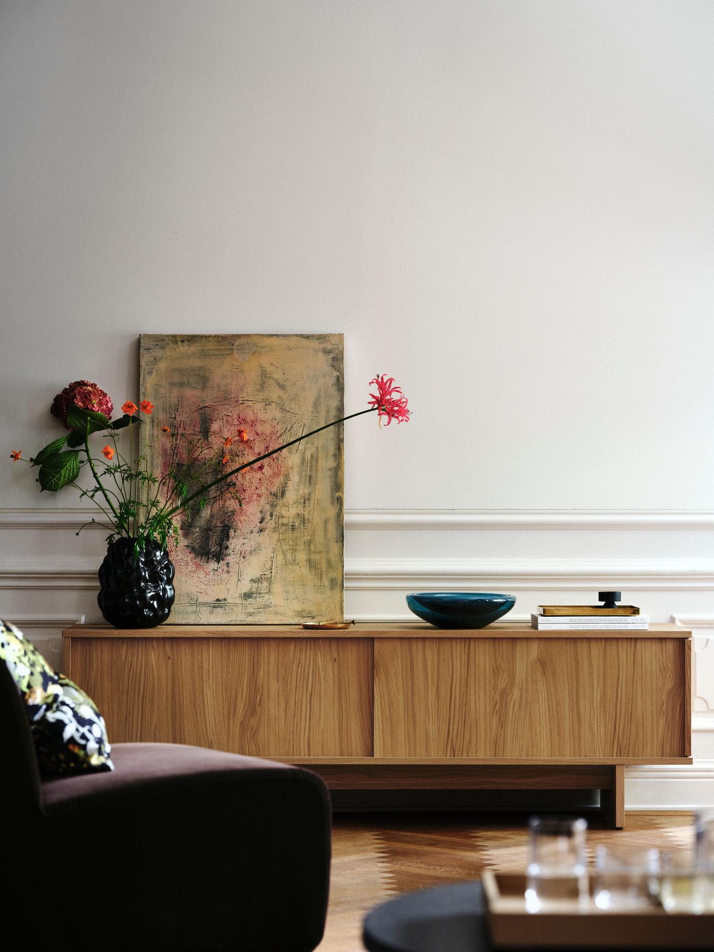 A low TV bench is standing towards a wall, with a painting and a vase with flower standing on top.