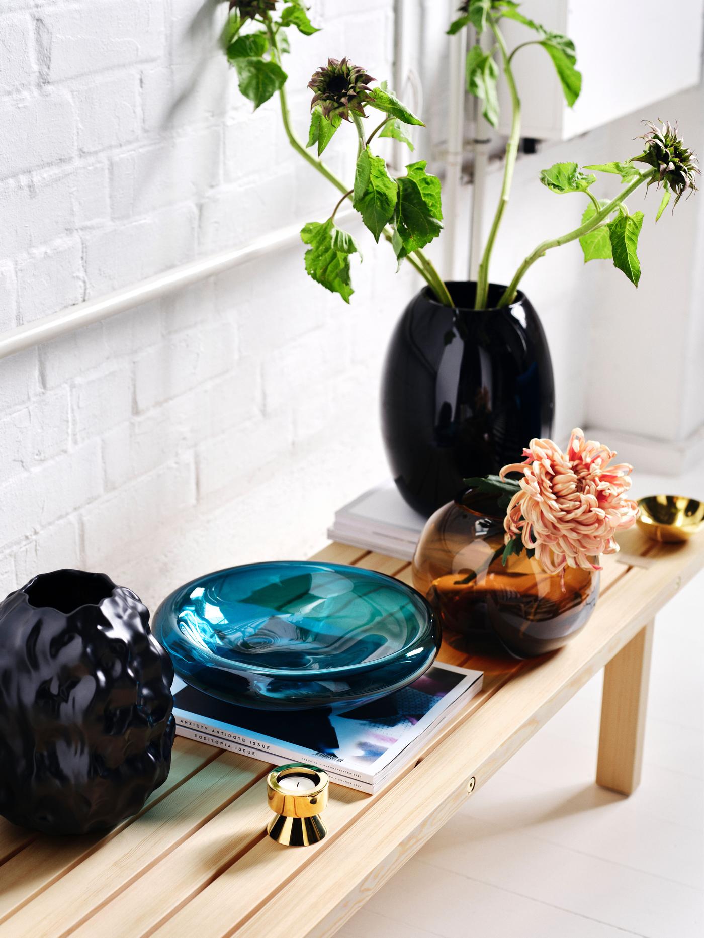 Four vases in black, blue, and light brown on a solid pine bench. Shot from the side, with green flowers in the large black vase.
