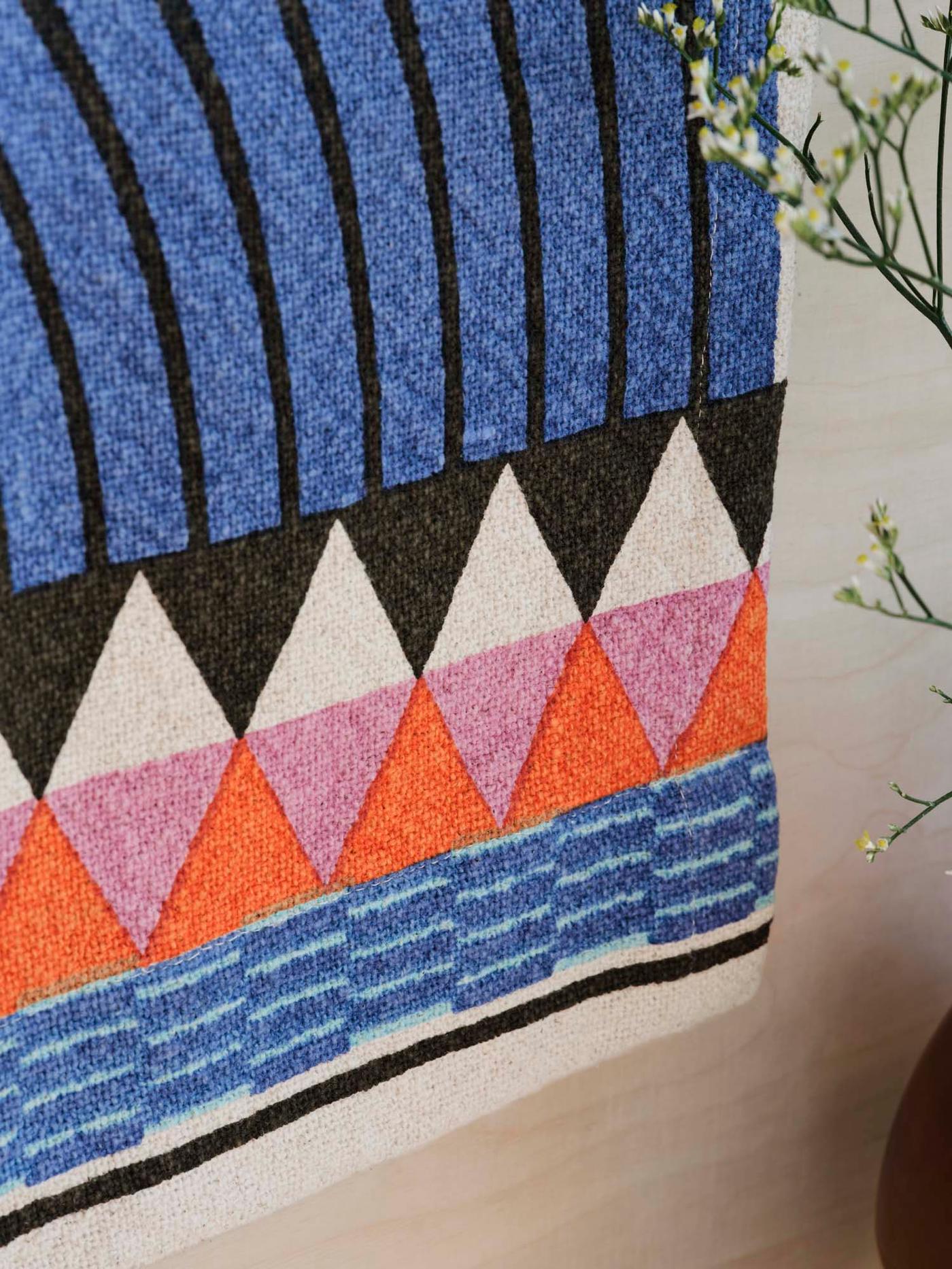 Close-up of a colourful hanging tapestry on a wood wall, next to a vase of flowers.