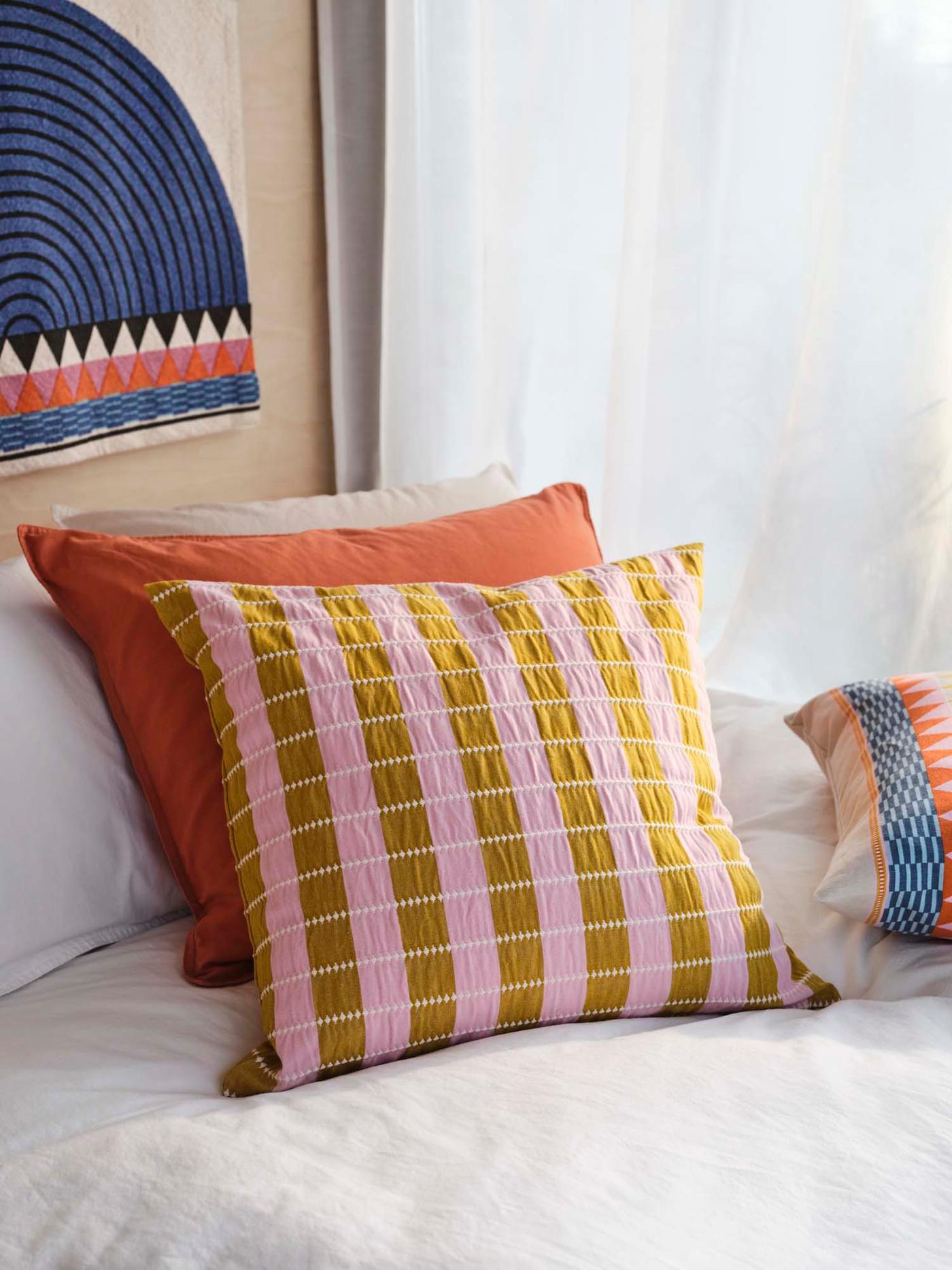 Three colourful cushions on a white bed, in front of a window.
