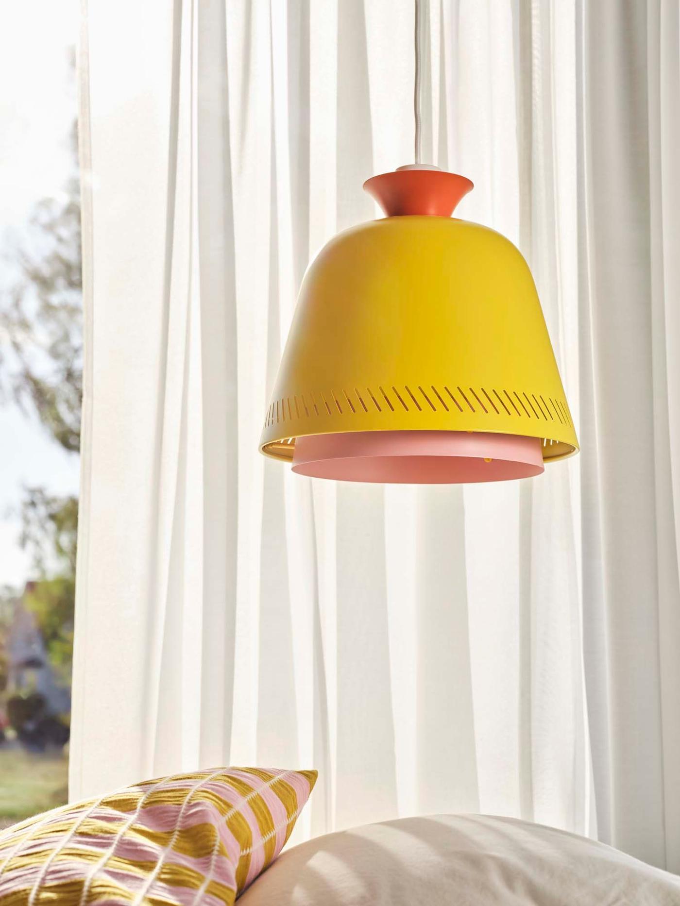 A yellow, pink and orange lamp hangs in front of a window and above a bed with cushions.