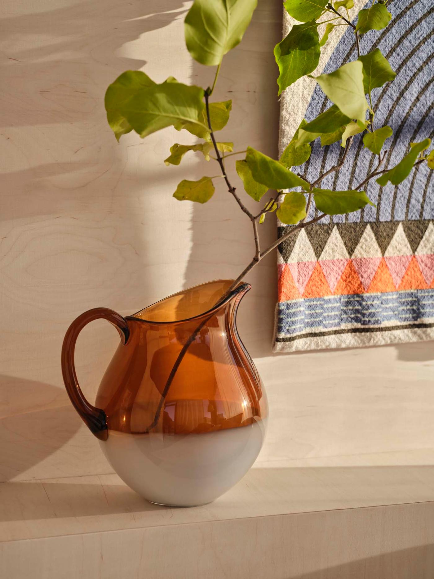 Shadows fall on an orange and white OMMJÄNGE jug with a green stem. It stands on a wood surface next to a wall hanging.