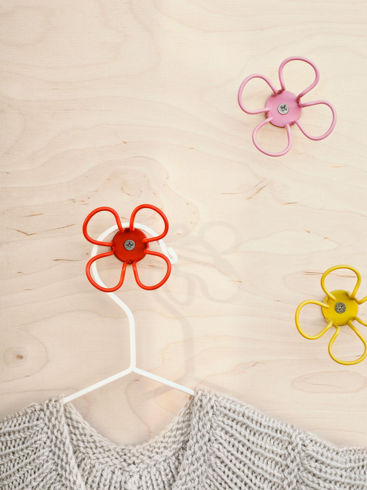 Three flower-shaped wall hooks in pink, yellow and red, with a hanger on the red hook.