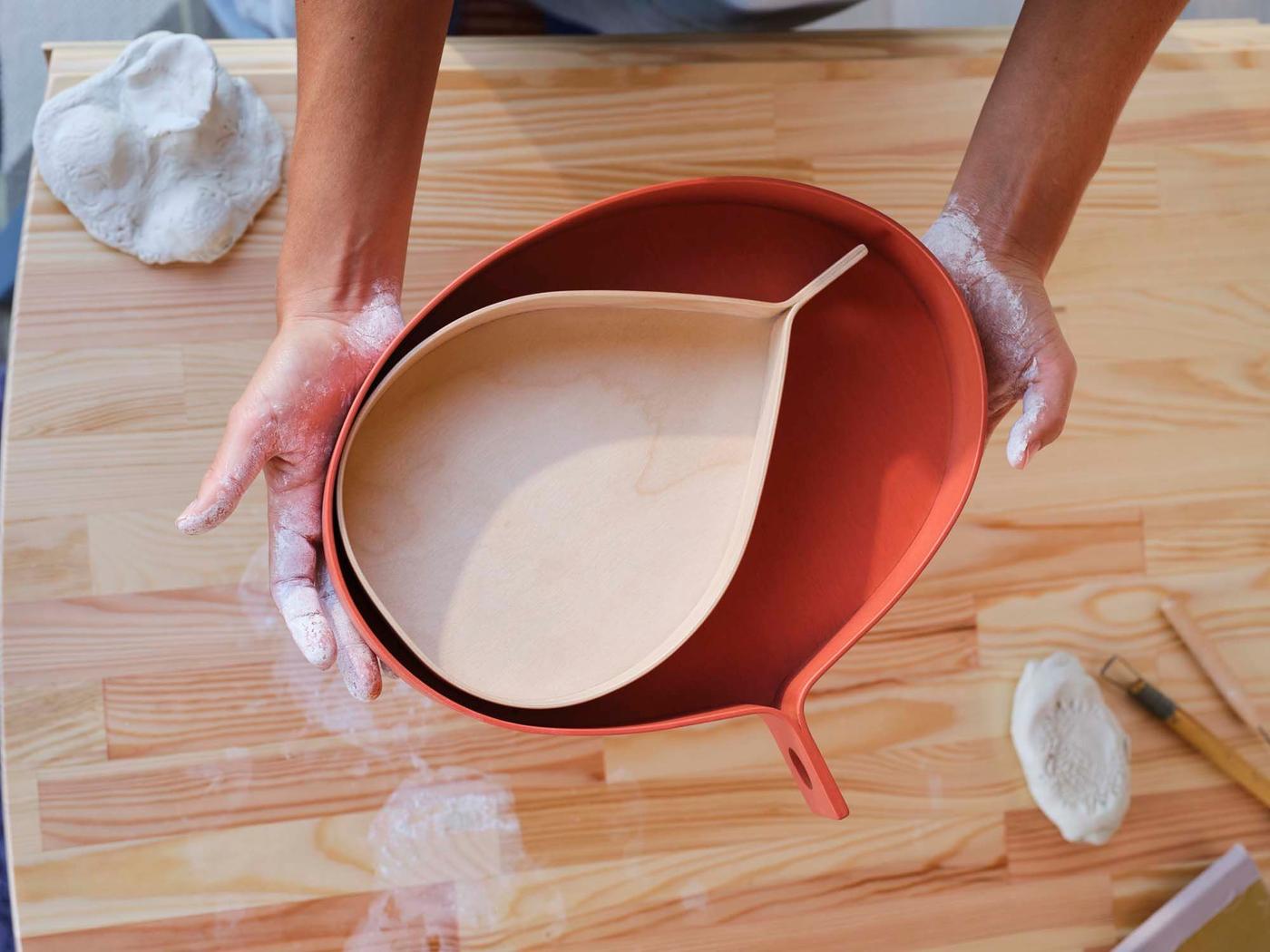 An aerial view of a pair of hands holding a pale wood tray inside a larger orange wood tray, above a wood surface.