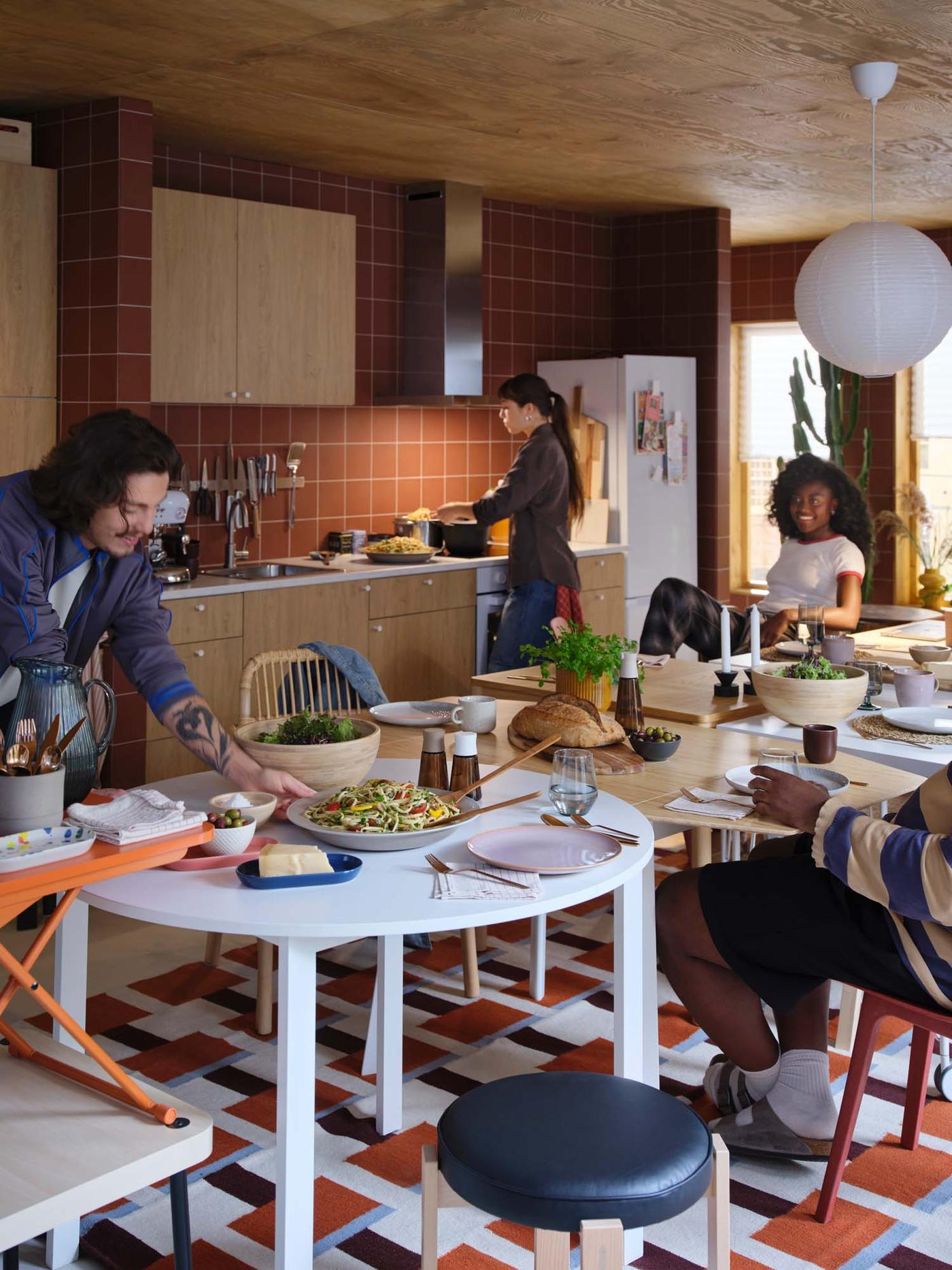 Three people sit and talk in a kitchen dining area, while one person prepares food at the worktop.