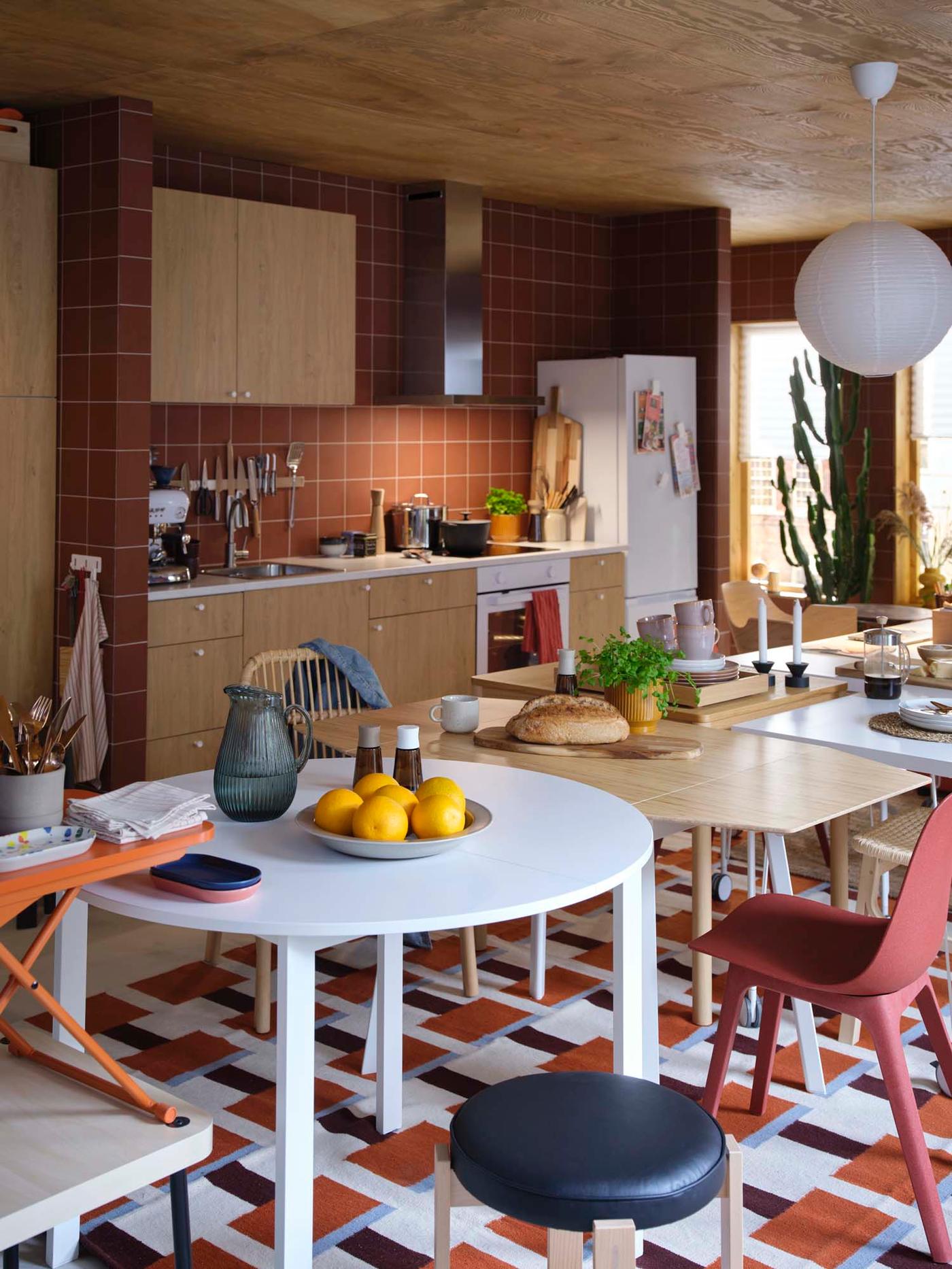 An oak-effect KNOXHULT kitchen with burnt orange tiles, set behind four mismatched tables pushed together and surrounded by mismatched chairs.