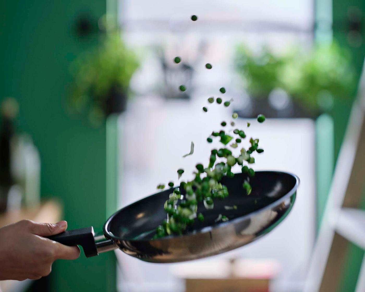 A person flips peas in a stainless steel MIDDAGSMAT frying pan.