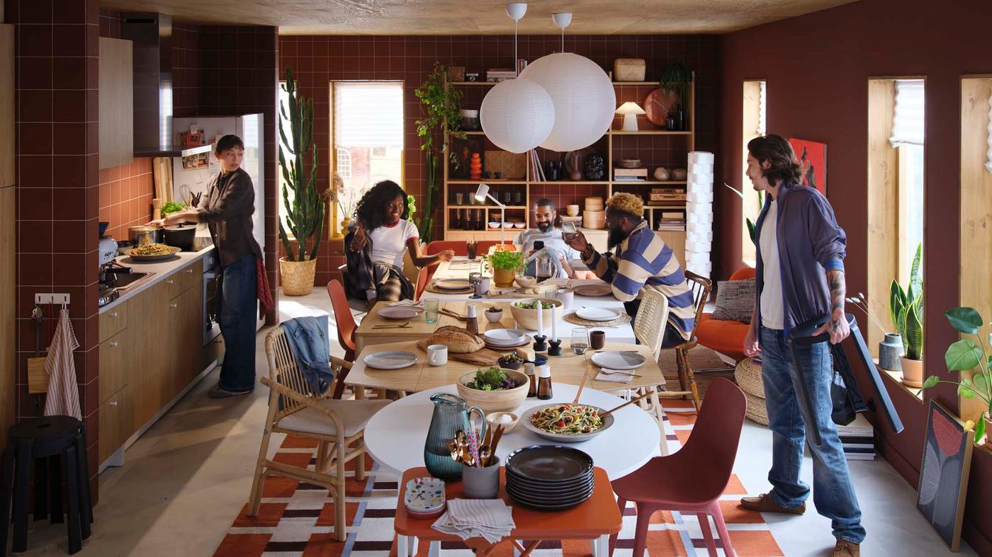 Four people gathered in a burnt-orange and light wood kitchen, sitting and standing around several tables pushed together with mismatched chairs.
