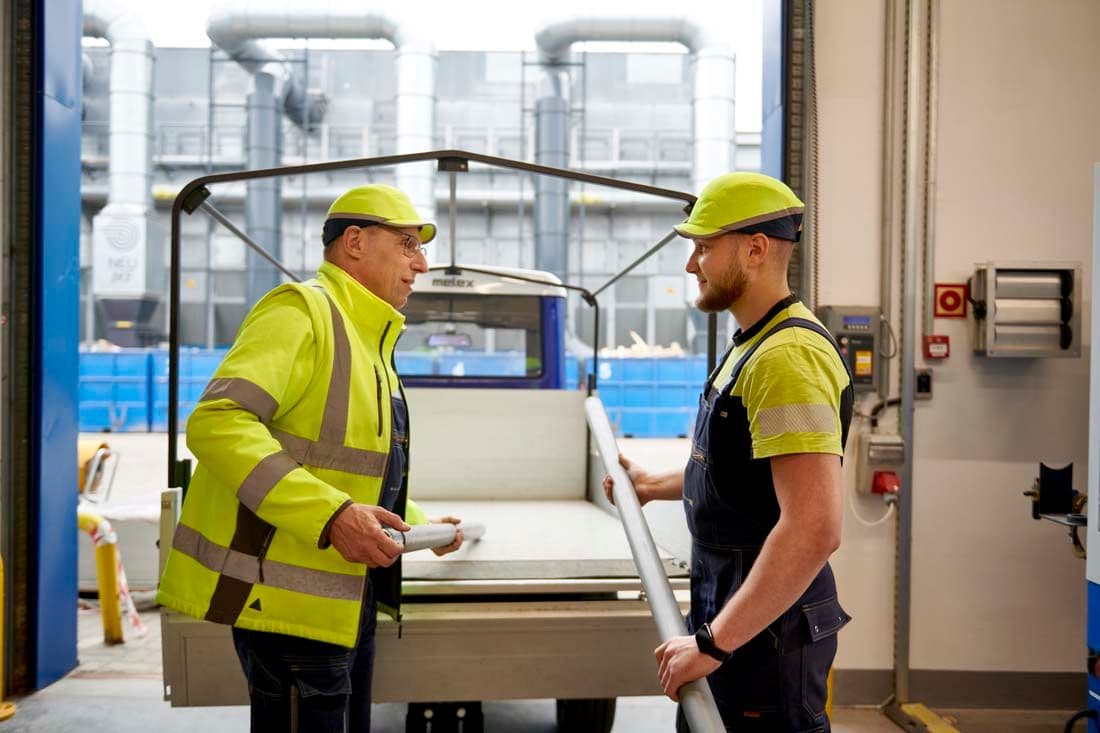 Two factory workers chat to one another while holding steel piping in a factory.