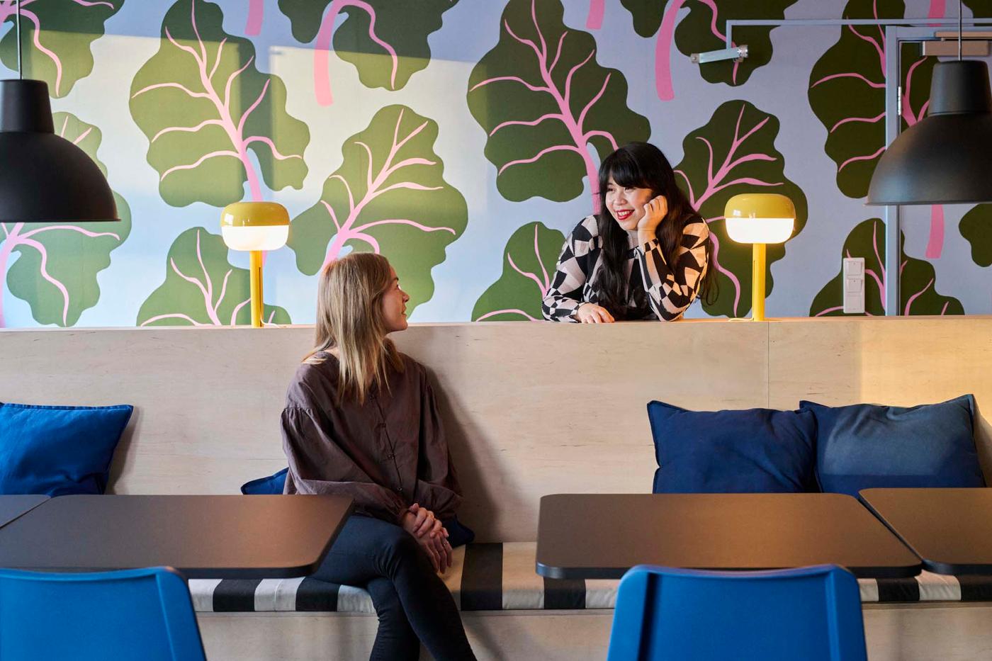 A person leans on the back of a wooden bench in a brightly coloured work environment. They are talking to a person sitting on the wooden bench in front.