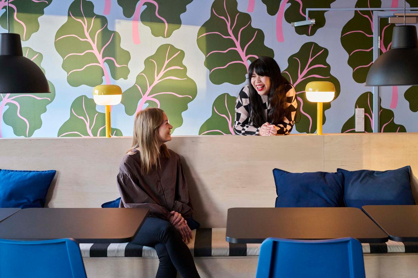 Two people are speaking. One leans on a wooden counter behind the other who sits and looks over their shoulder to speak.