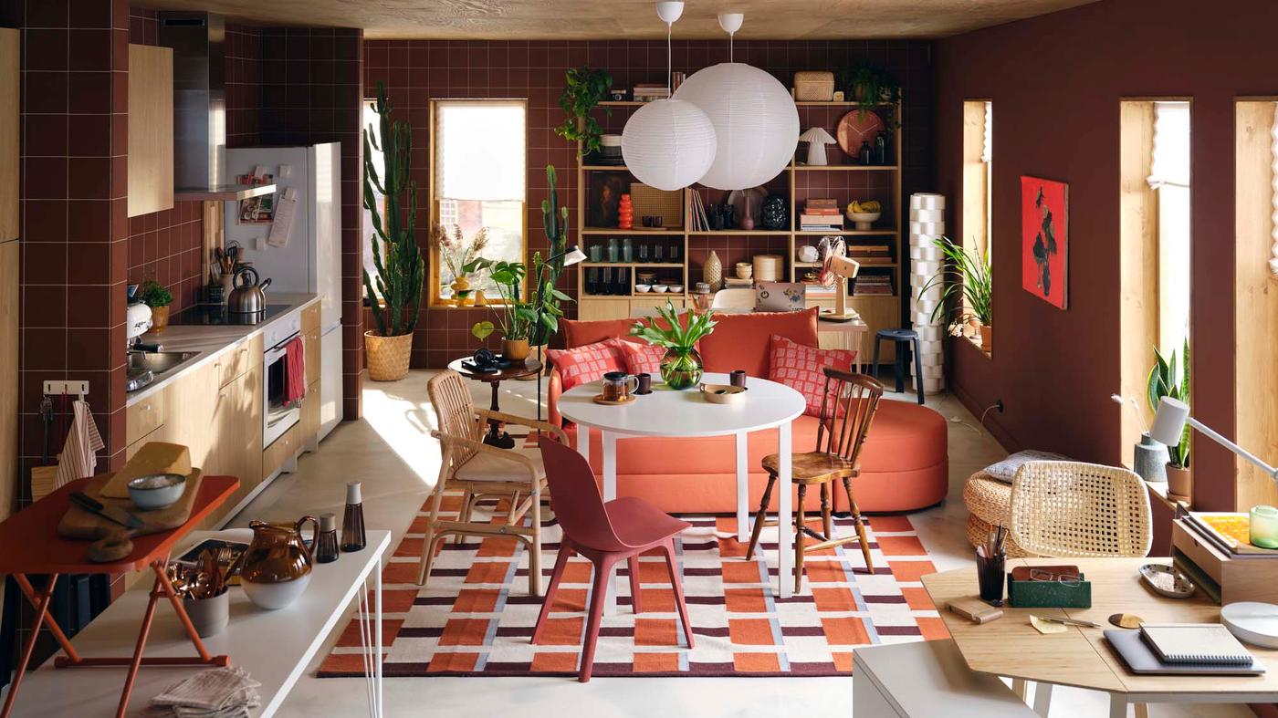 An open-plan room with a LINDÅKRA sofa-bed used as seating at a round table beside an oak, white and burnt orange kitchen.