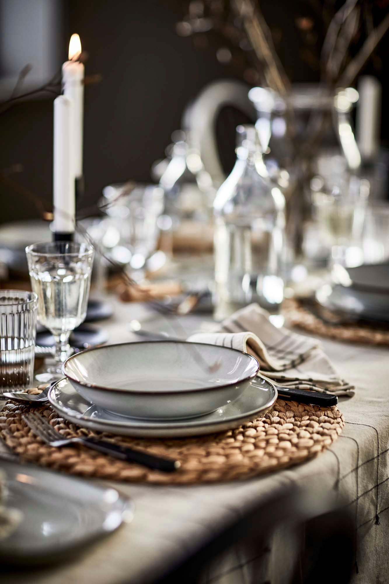A calm table setting with a grey GLADELIG plate and bowl on a woven placemat, surrounded by glassware on a light grey tablecloth.