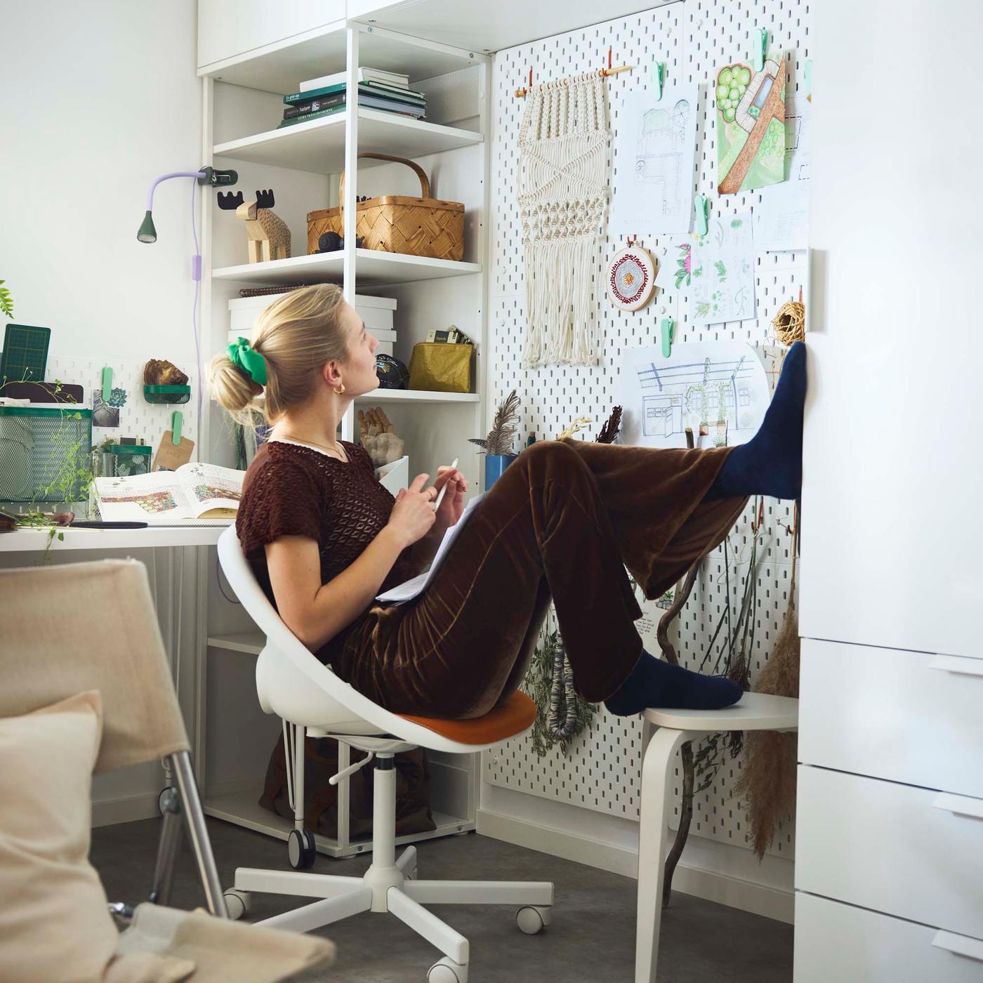 A college student wearing a brown t-shirt and brown pants sits in a white rolling desk chair. They have their feet up on the white desk in front of them and are looking toward papers hanging on a white pegboard.