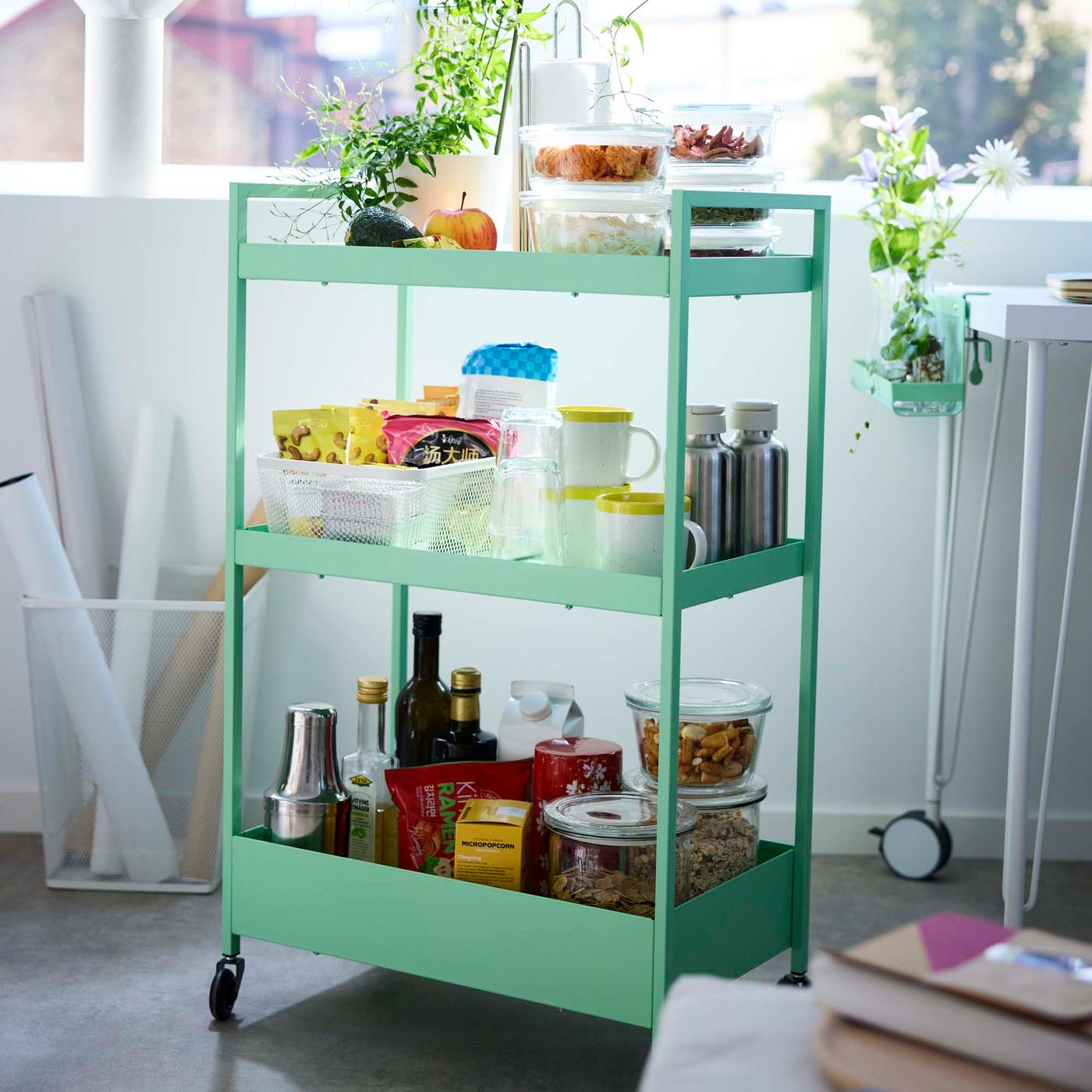 A light green rolling kitchen trolley with three shelves hold items for making snacks such as coffee cups, tea and food storage containers.