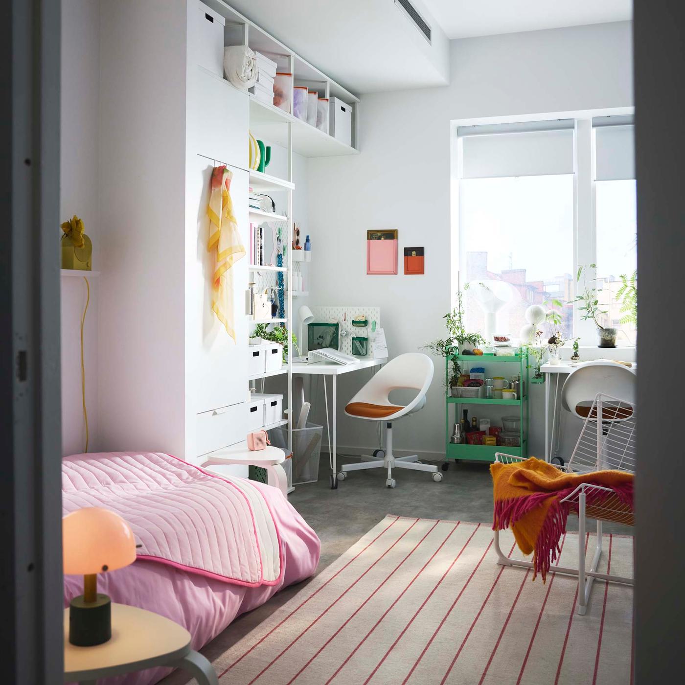A view of a bedroom with a striped rug. A desk area is in the background, positioned along a wall next to a white wardrobe. A bed in the foreground is dressed with a pink duvet cover.