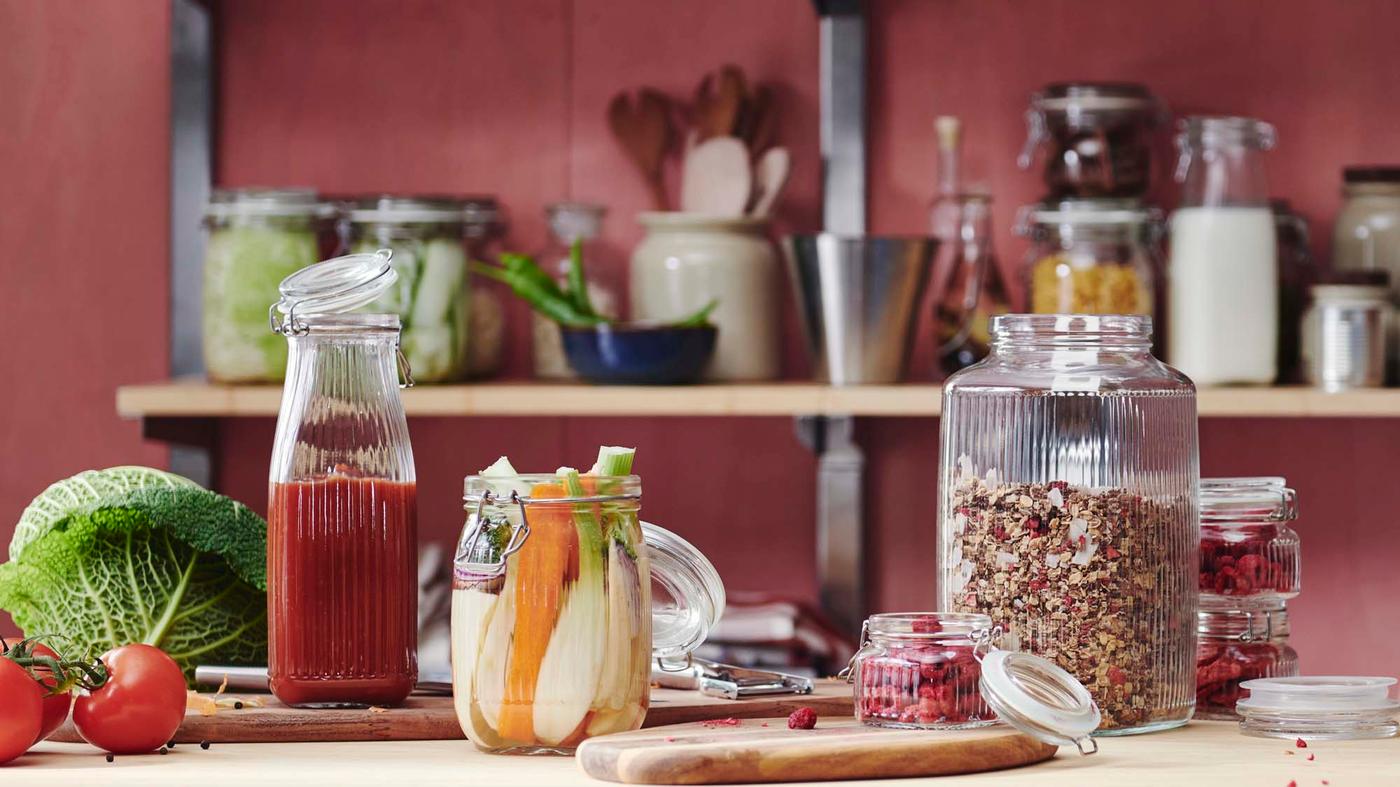 A bottle filled with blended tomato, and jars filled with vegetables, raspberries and granola sit on a wooden table, with jars in the background.