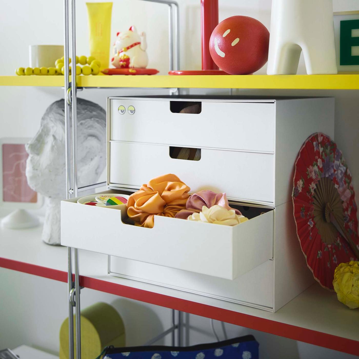 A white storage box with four drawers sits on the shellf of a storage unit. One drawer is pulled open and a collection of hair scrunchies are inside.