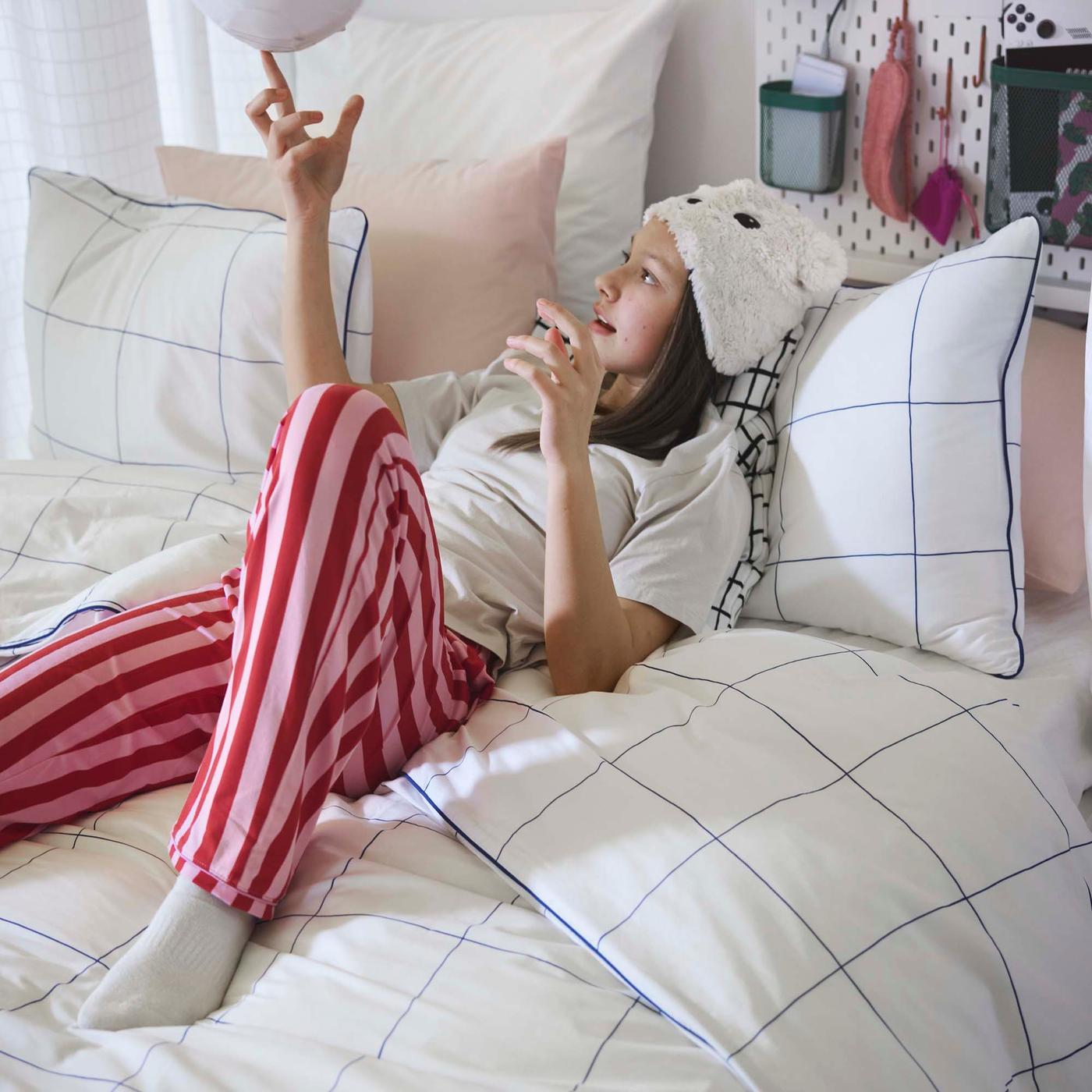 A teenager in a white t-shirt, white knit hat and pink and red striped pajama pants reclines on a bed. The bedding is white with a black lined grid pattern.