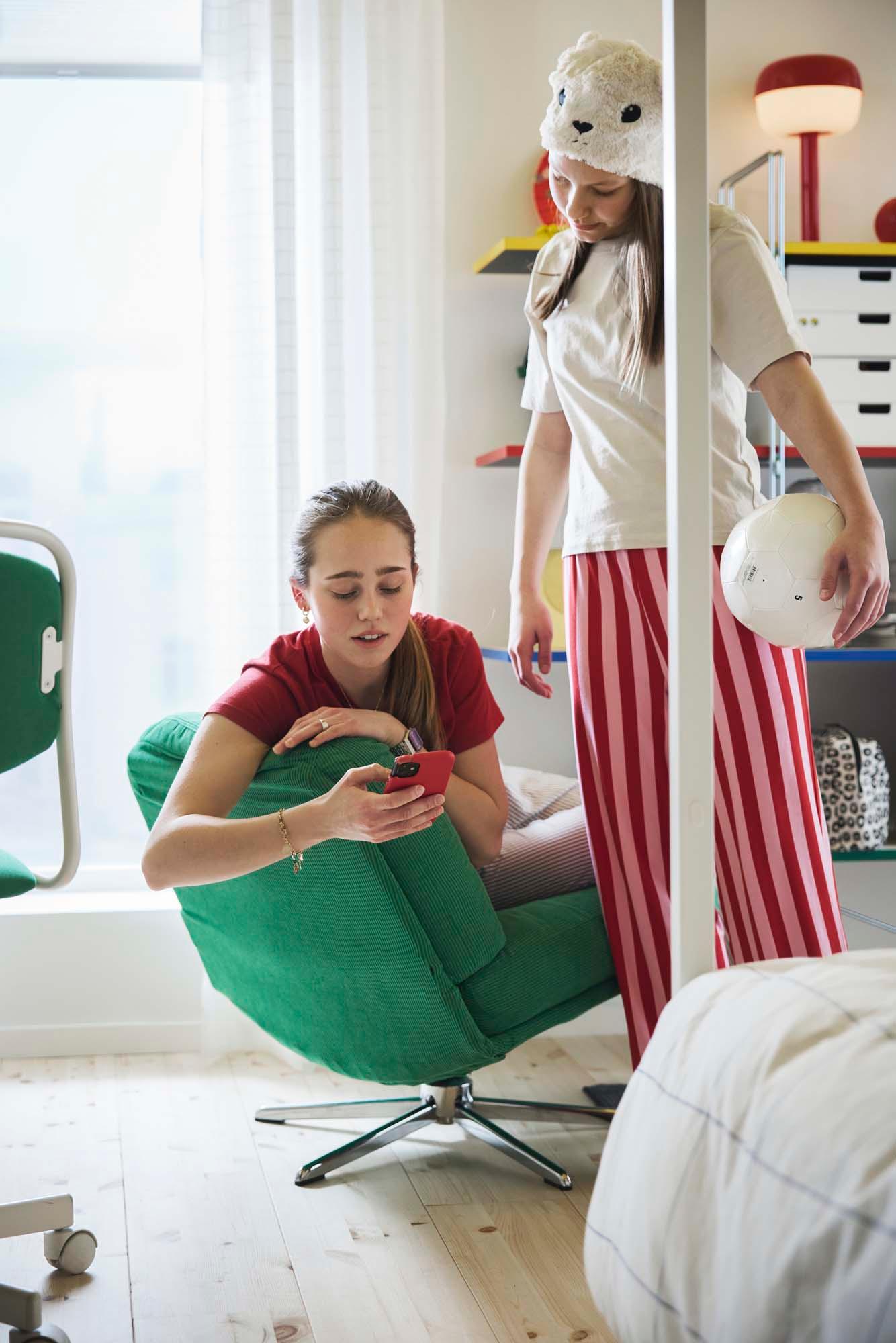 Two teenagers are talking together. One is sitting in a bright green low lounge chair. The other is standing to the side, wearing striped pajamas and holding a white ball.
