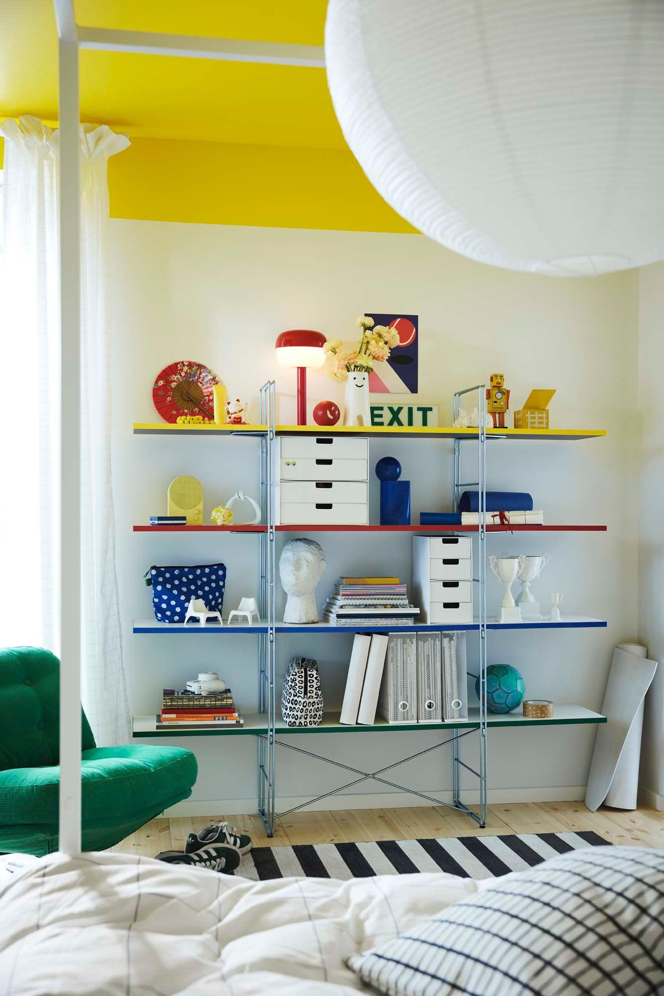A large storage unit has four shelves, each with a primary colour decorating the edge; bright red, blue, green and yellow. A red lamp, books and white storage boxes are on the shelves, along with other colourful decorative items.s.