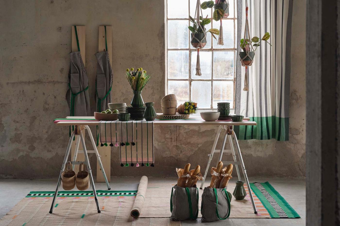 A trestle table with vases, bowls and plates, with rugs below and plants at the window.